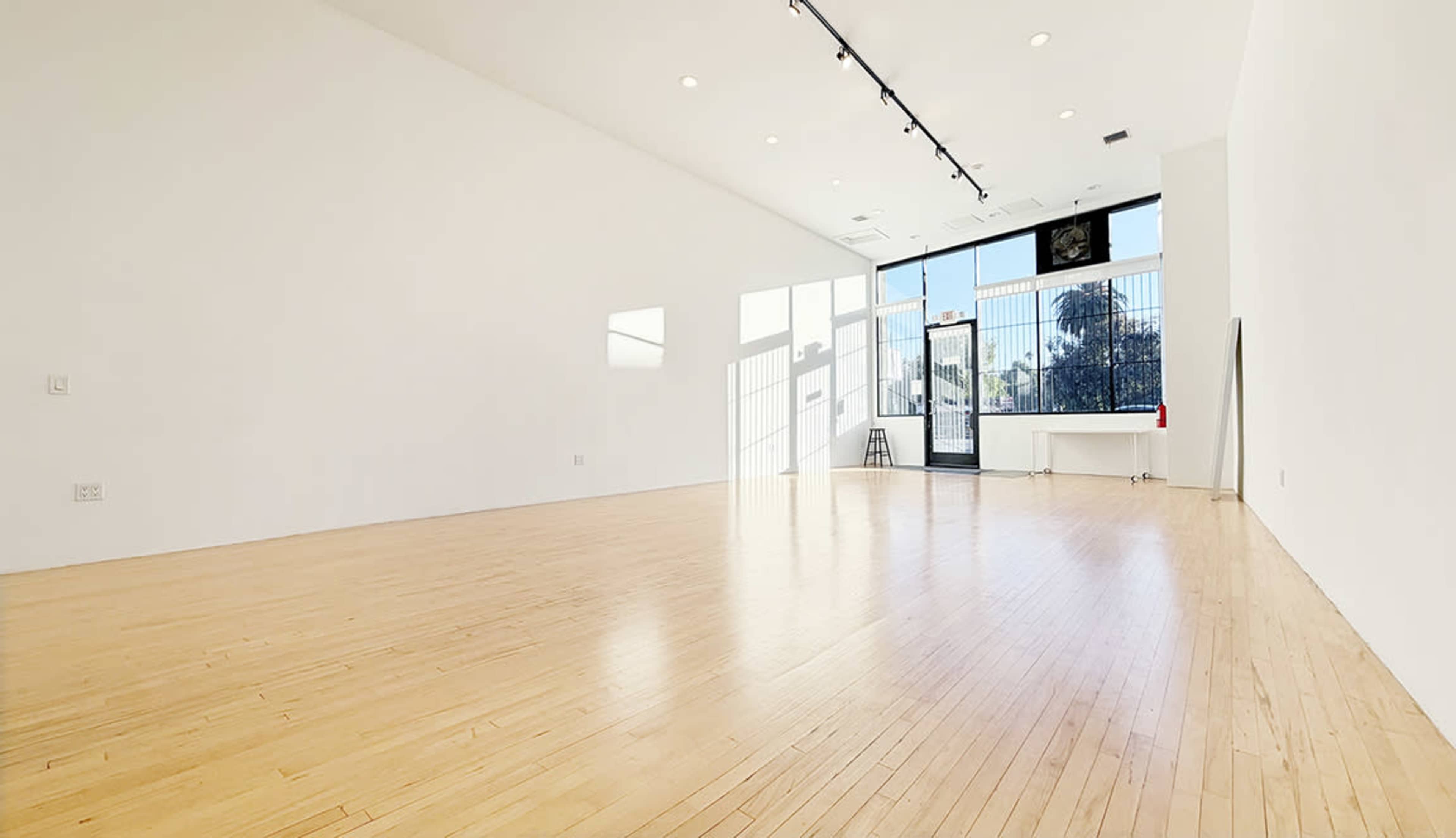 The image shows a spacious, well-lit interior with wooden flooring, large windows, and minimal furniture.