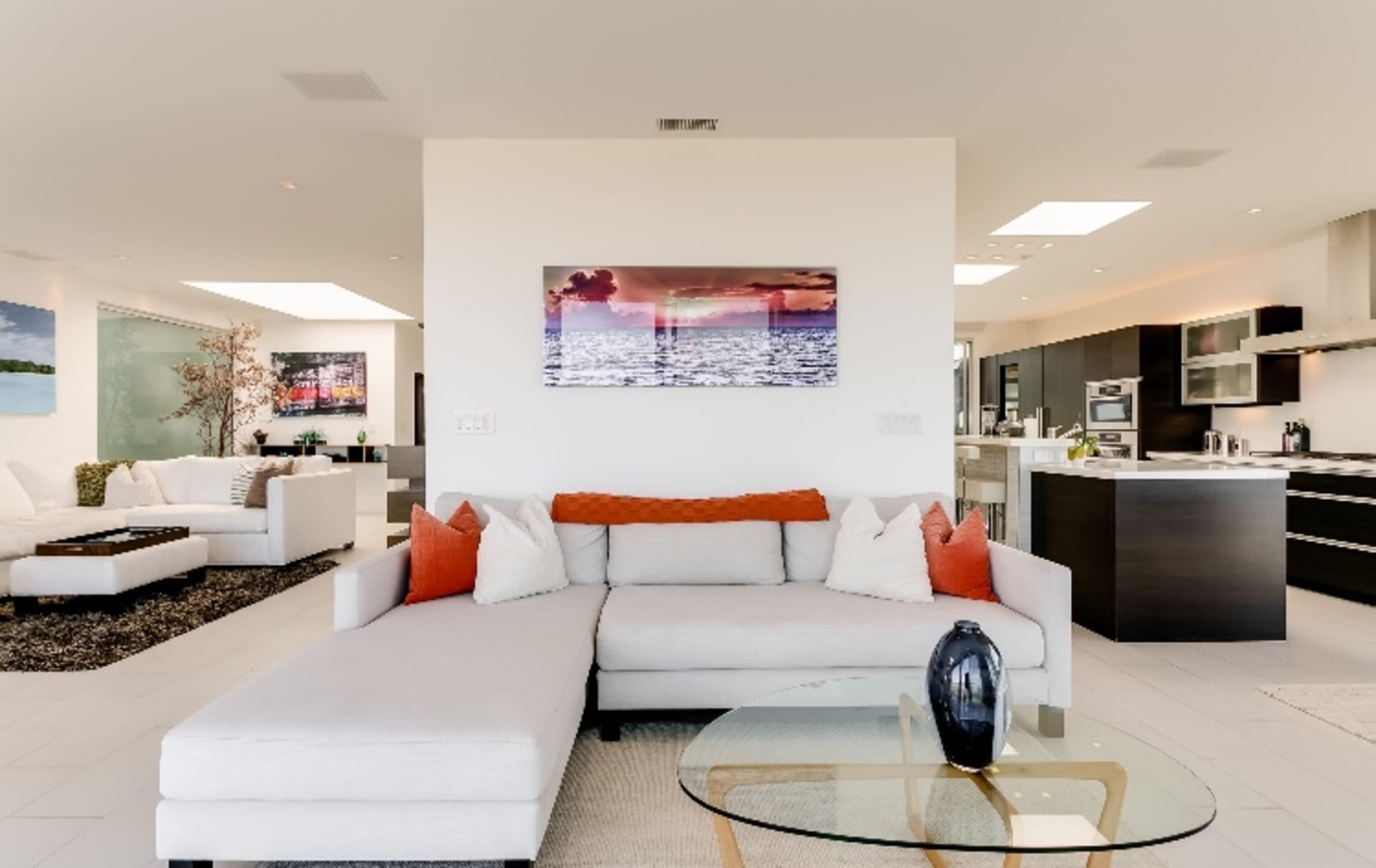 A modern living room features a light-colored sectional sofa with orange accent pillows, a glass coffee table, and a wall-mounted artwork, with a kitchen and another seating area visible in the background.