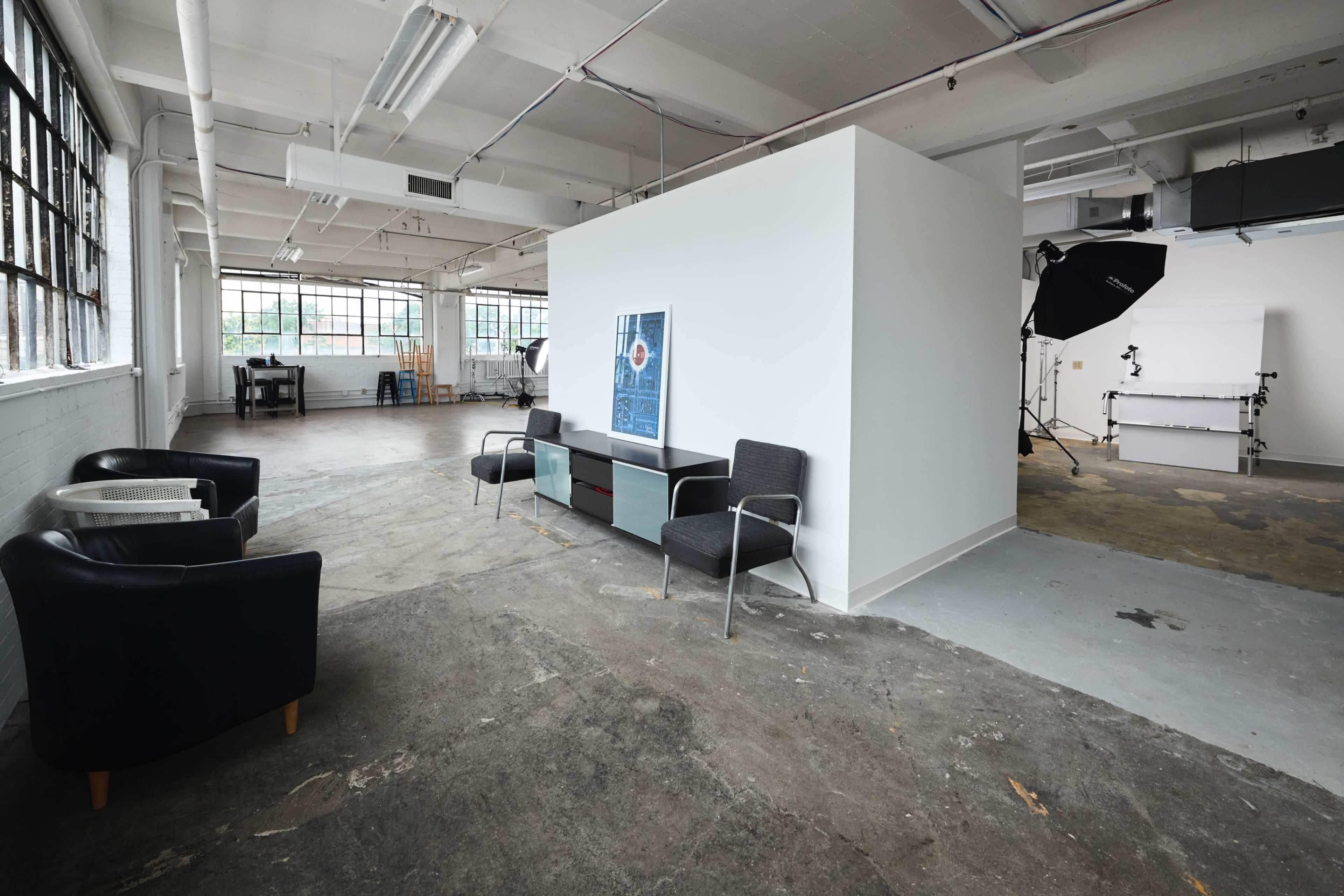 A spacious, industrial-style room features a dividing wall with a framed artwork and black chairs, along with natural light streaming through large windows.