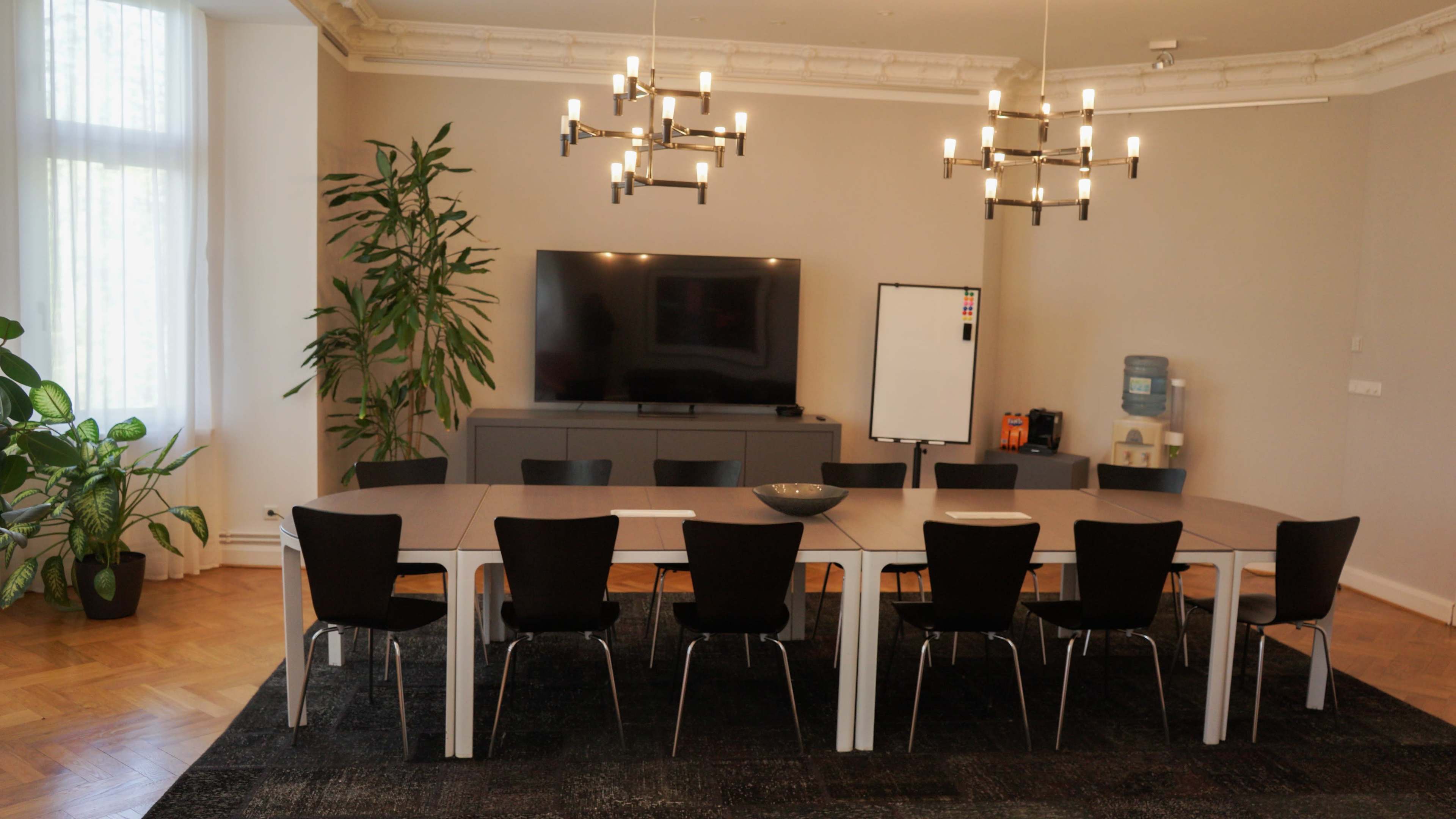 The image shows a modern conference room with a long table surrounded by black chairs, a large TV on the wall, and a whiteboard.