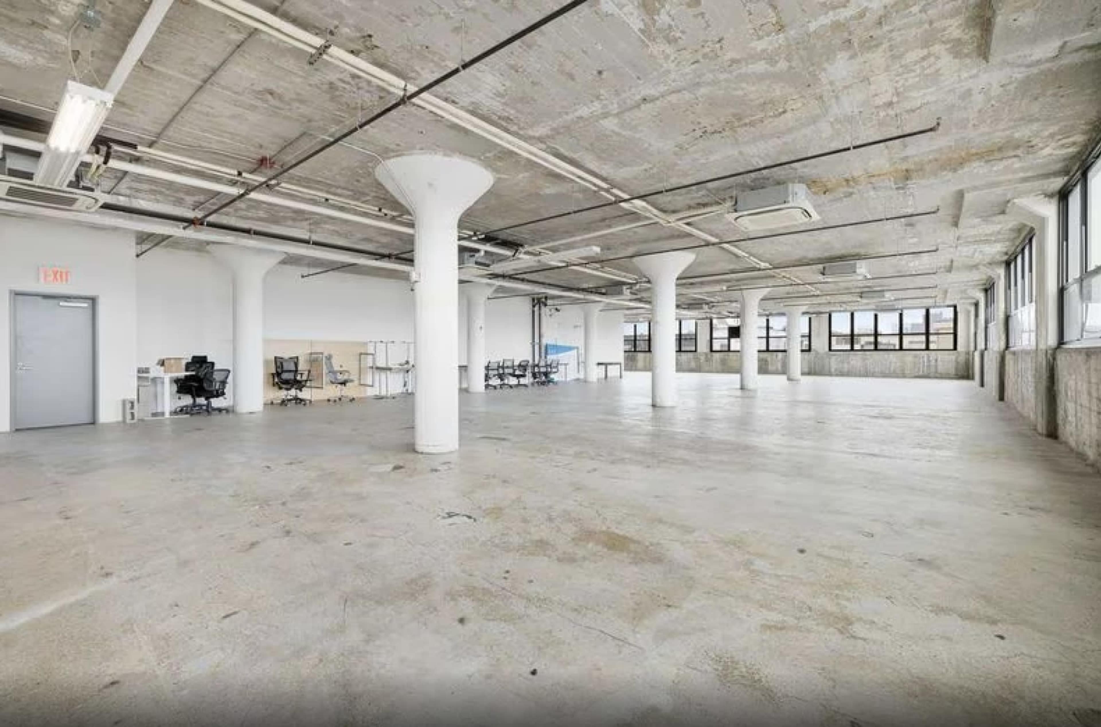 The image shows a spacious, empty industrial-style office interior with concrete floors and large windows.
