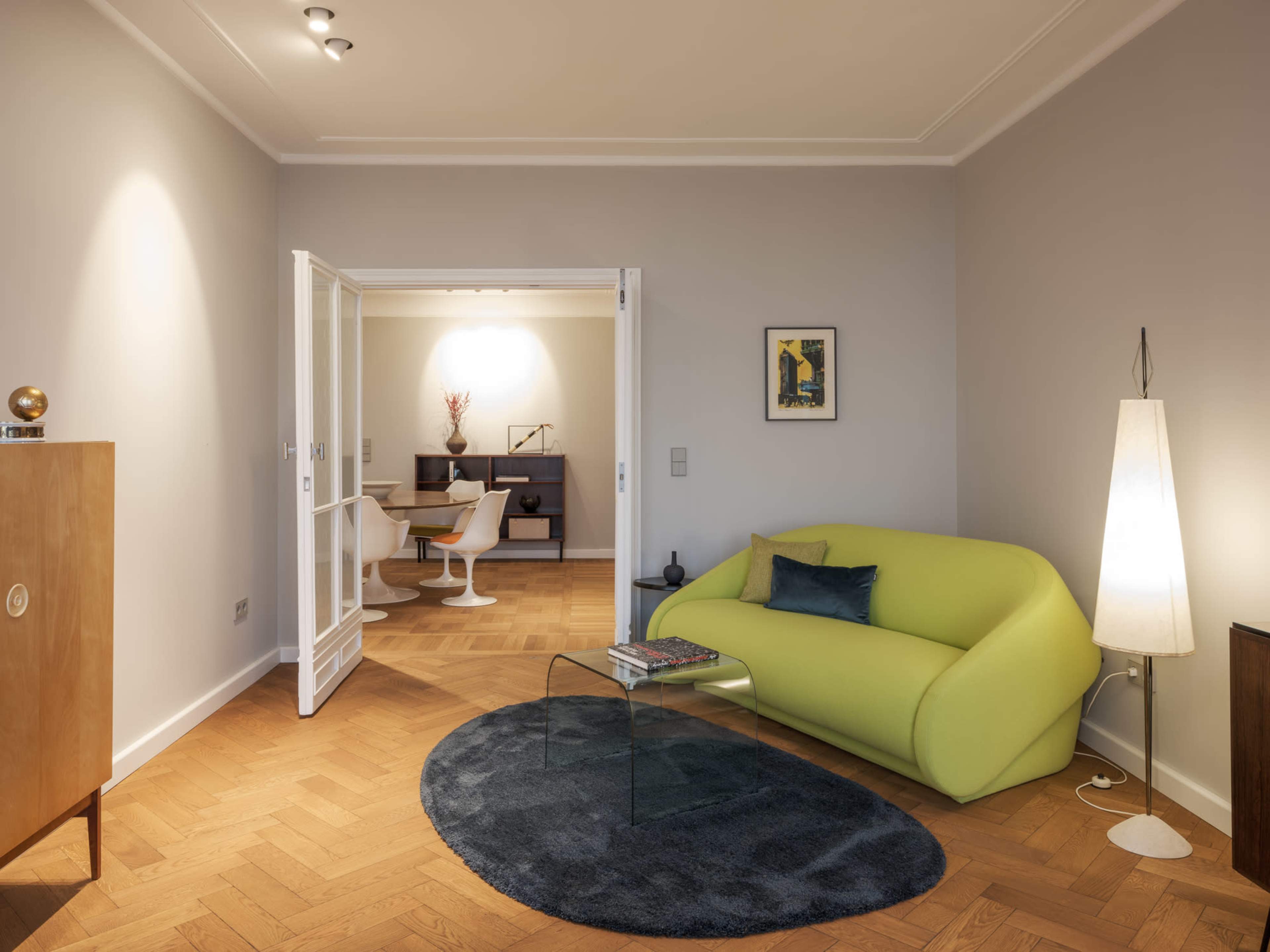 A modern living room with a green sofa, glass coffee table, and wooden flooring, leading to another room through open doors.
