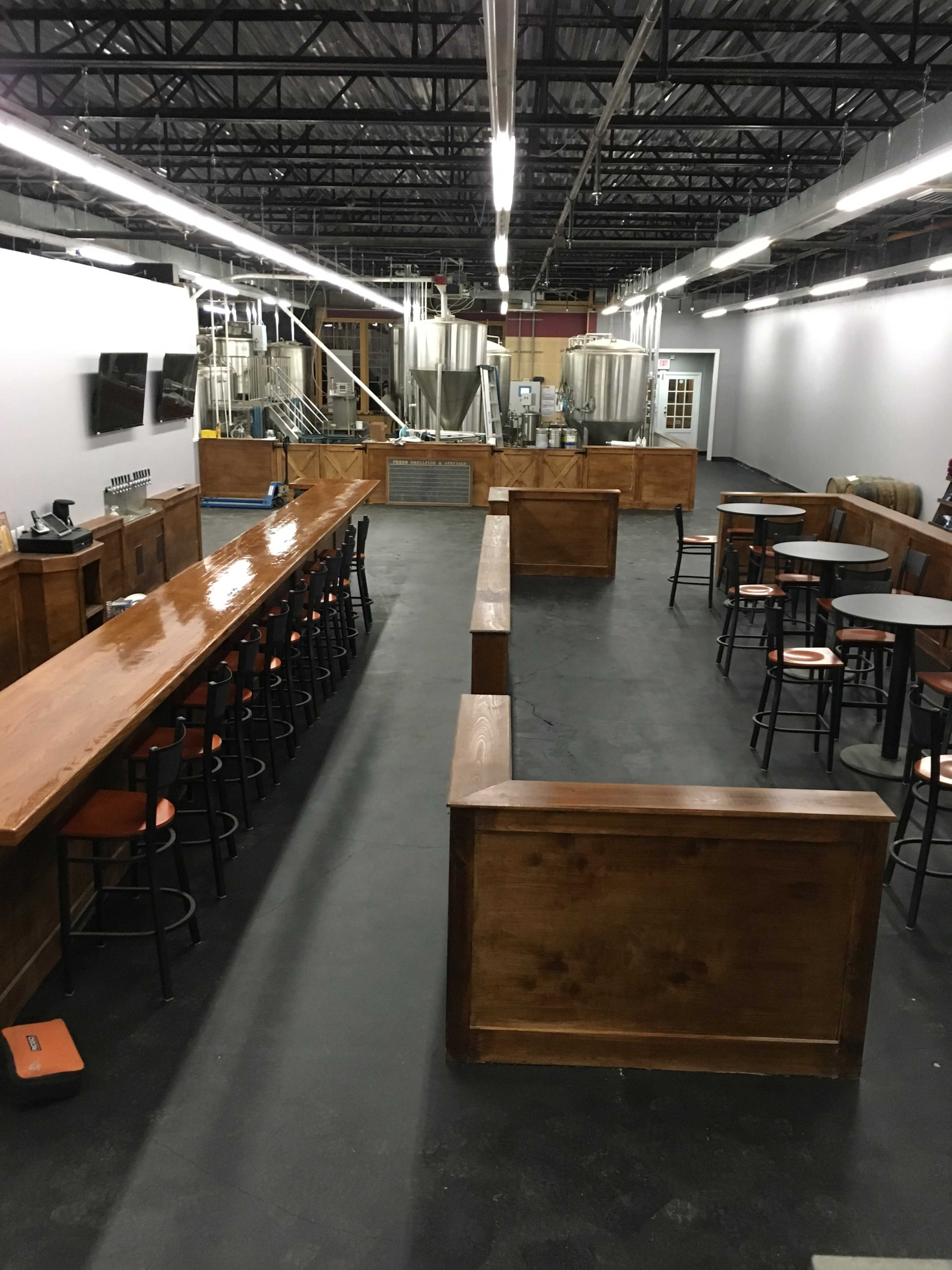The image shows a spacious brewery interior with a wooden bar, seating areas, and large brewing tanks in the background.