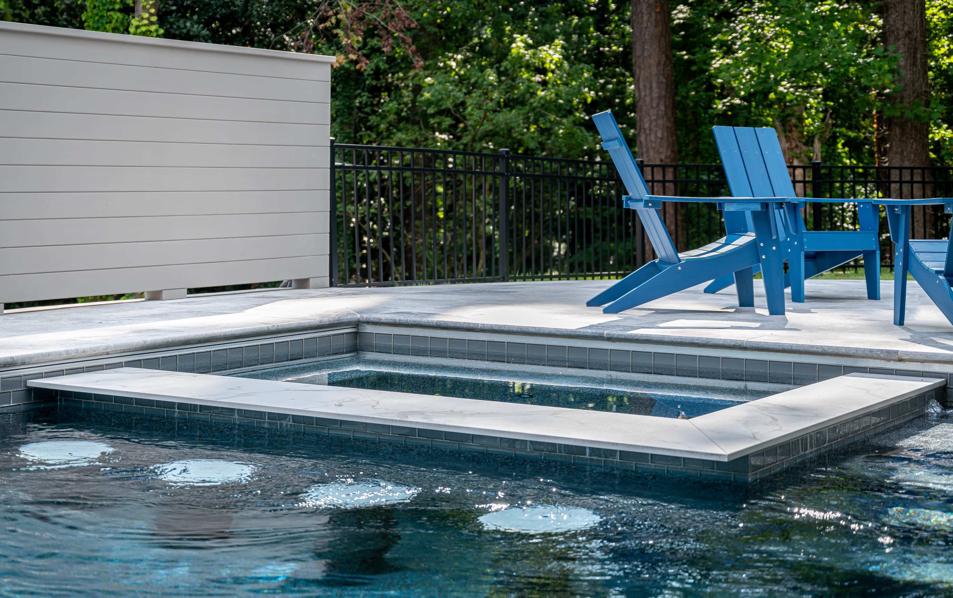 The image shows a backyard pool with a raised spa area and blue adirondack chairs nearby, surrounded by greenery.