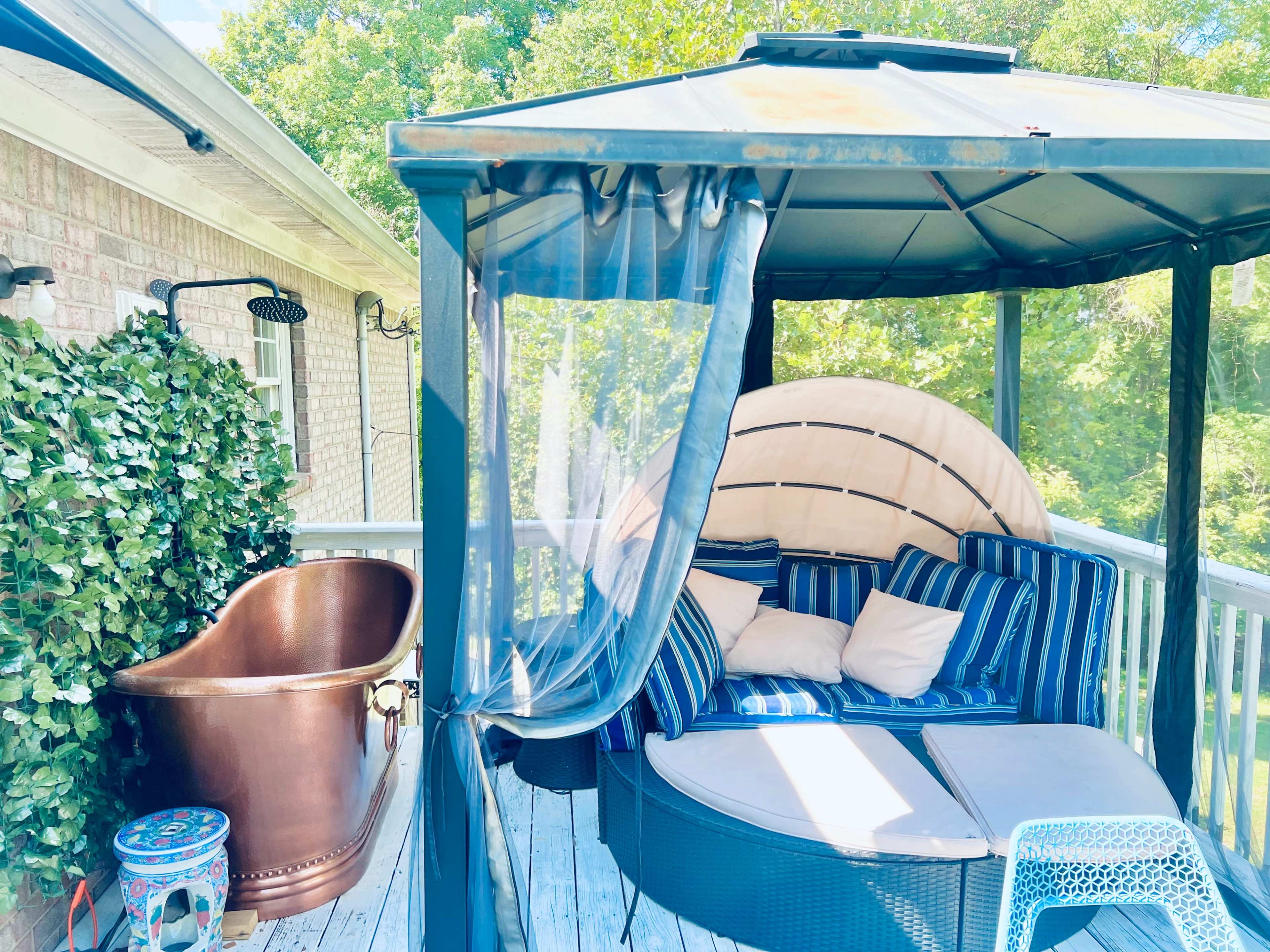 A gazebo with a round, cushioned seating area and a copper bathtub is set on a wooden deck surrounded by greenery.