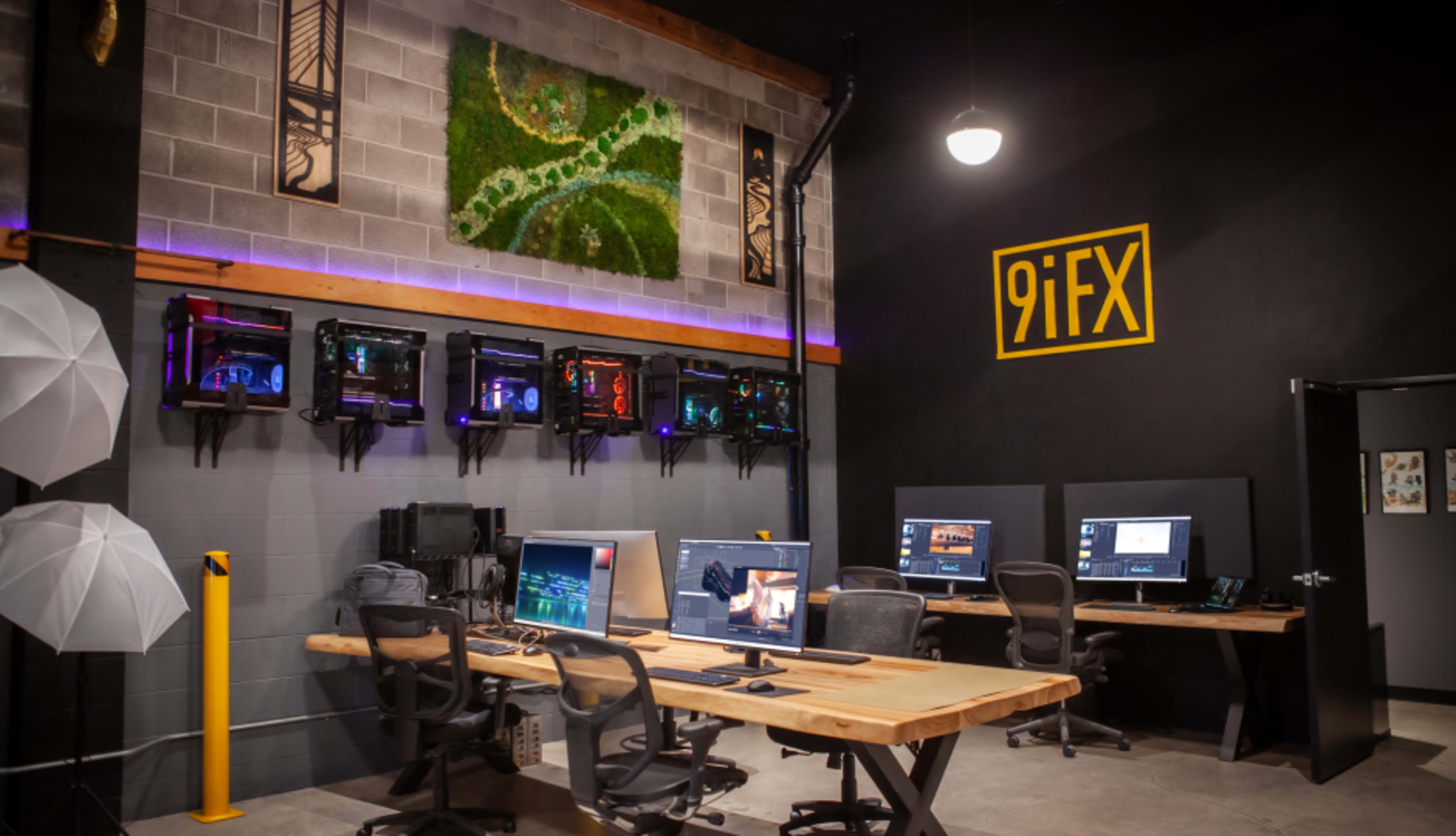 The image shows a modern workspace featuring multiple computer stations, a large wooden table, colorful wall art, and illuminated computer setups.