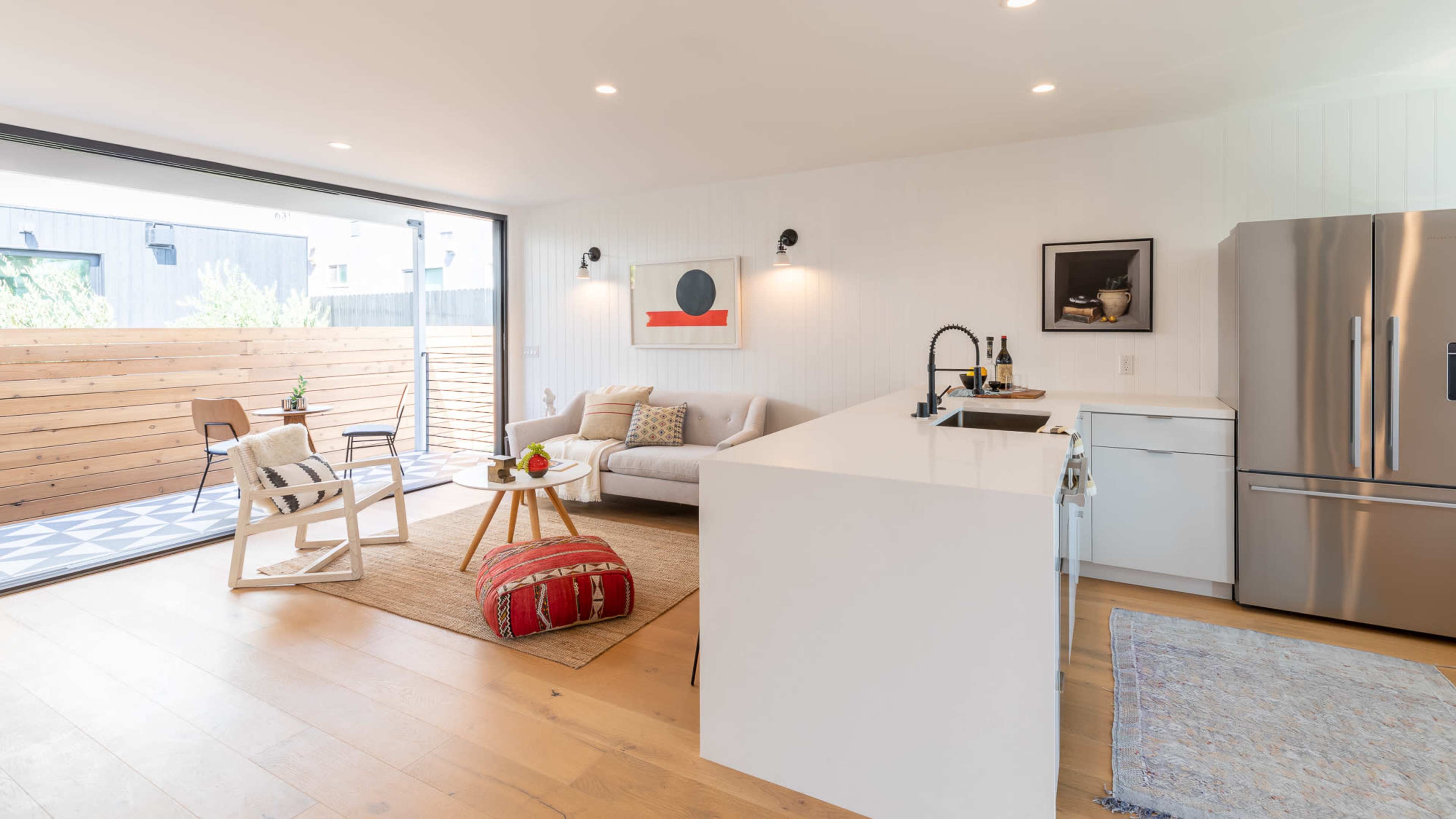 A modern kitchen and living room area features a white island, stainless steel appliances, a sofa, and a sliding glass door opening to an outdoor patio.