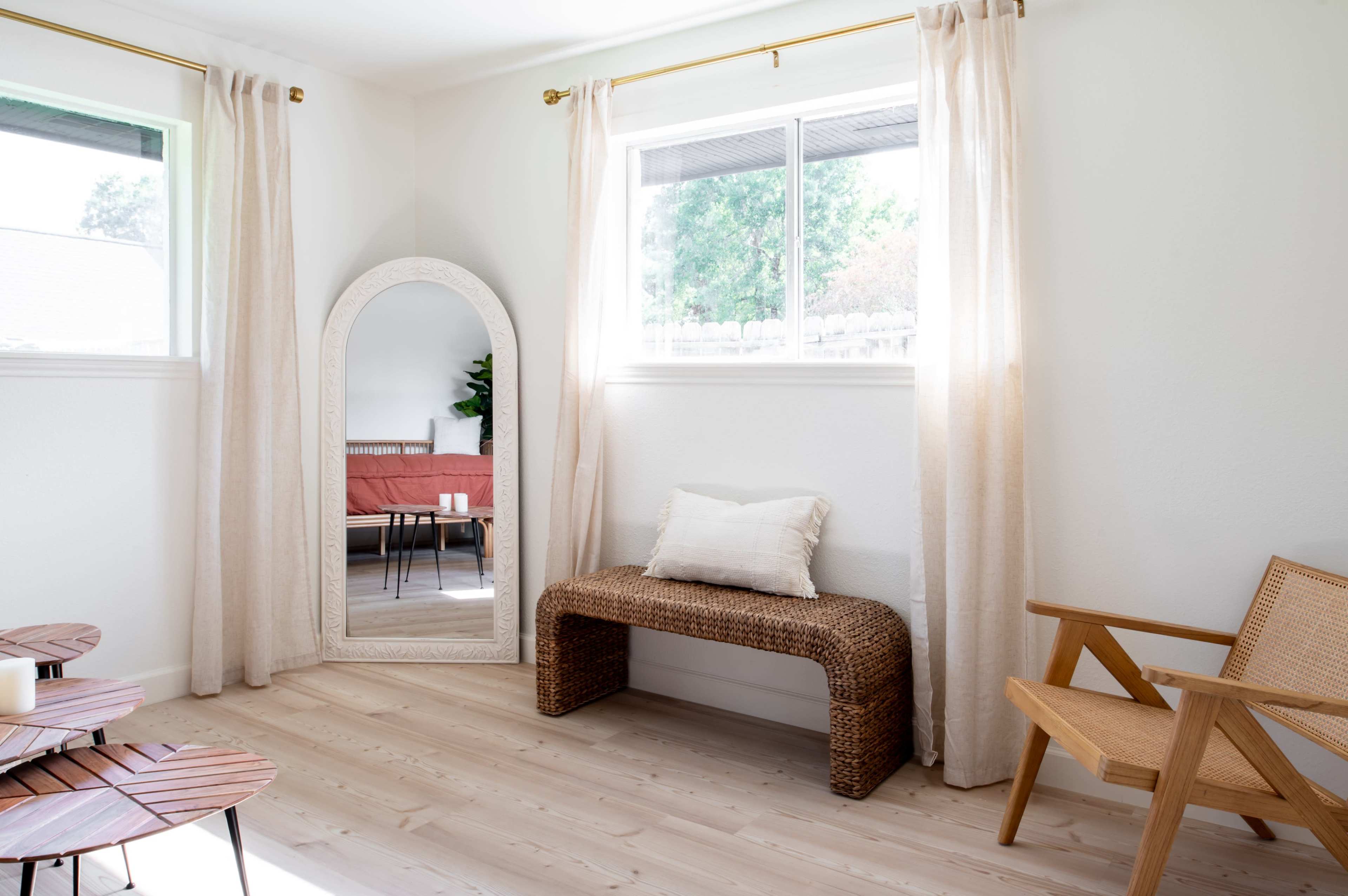 A room features a woven bench and a wooden chair beside a large mirror, with natural light coming through the windows.