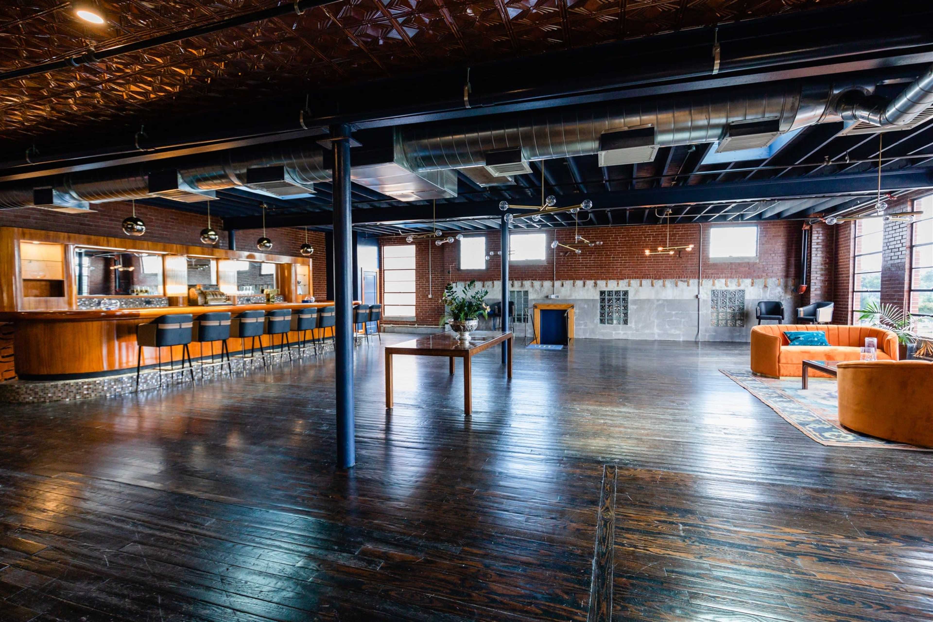 A spacious industrial-style room with wooden flooring, exposed brick walls, and a modern kitchen area featuring a long bar with stools.