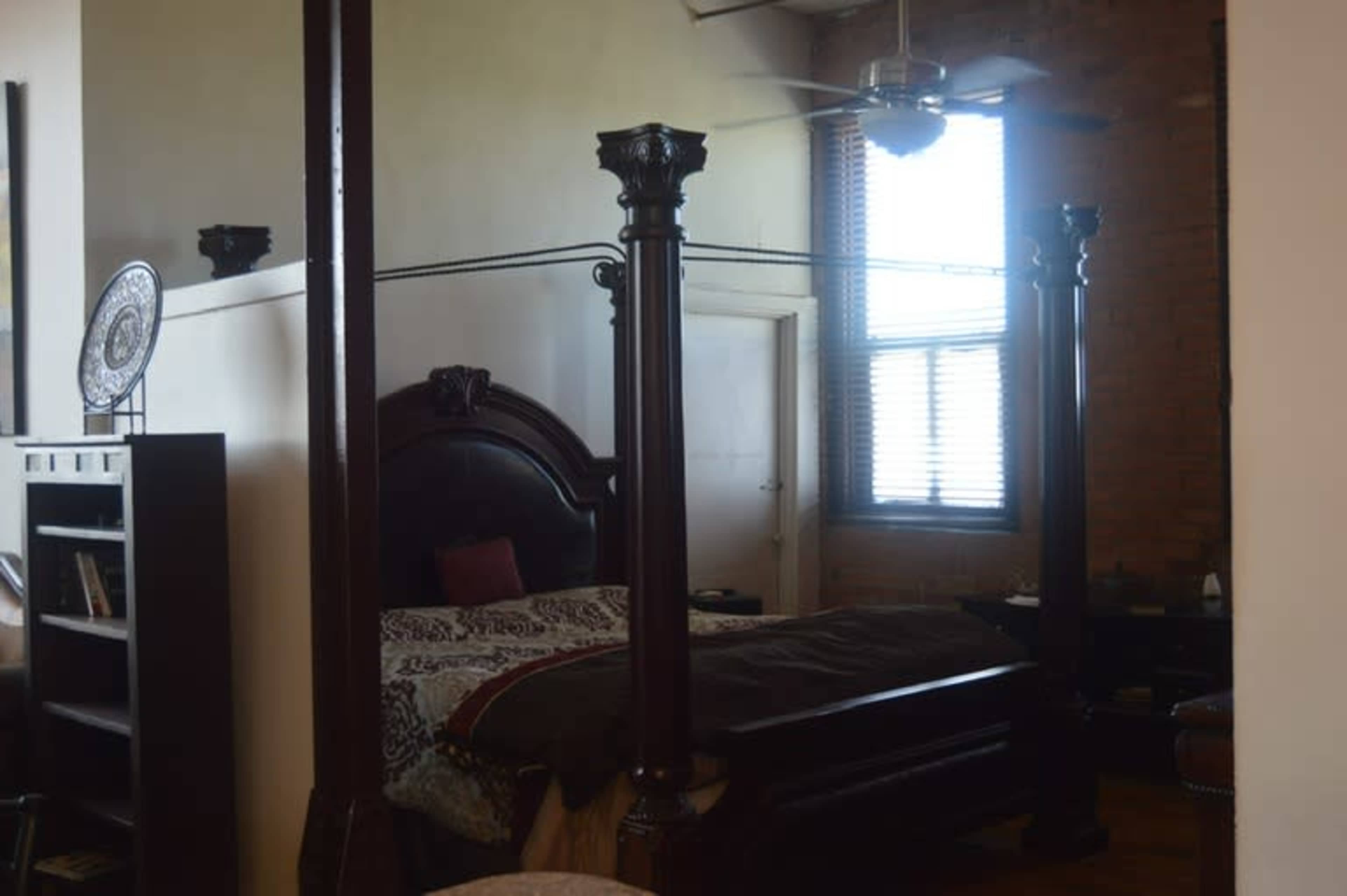 A bedroom featuring a four-poster bed, a ceiling fan, and windows with blinds, alongside wooden furniture.