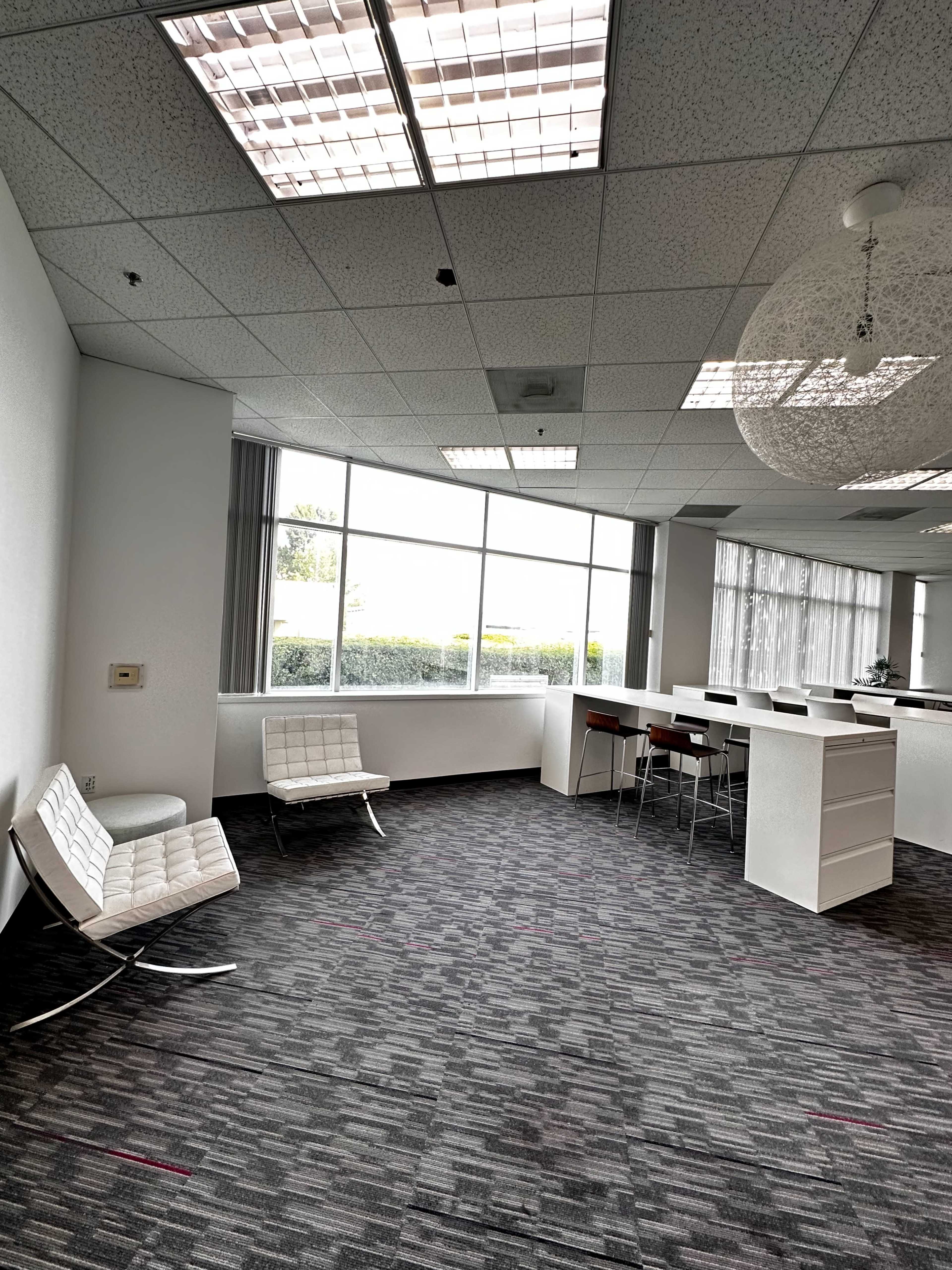 A modern office space featuring two white chairs, a bar-height table with stools, and large windows letting in natural light.