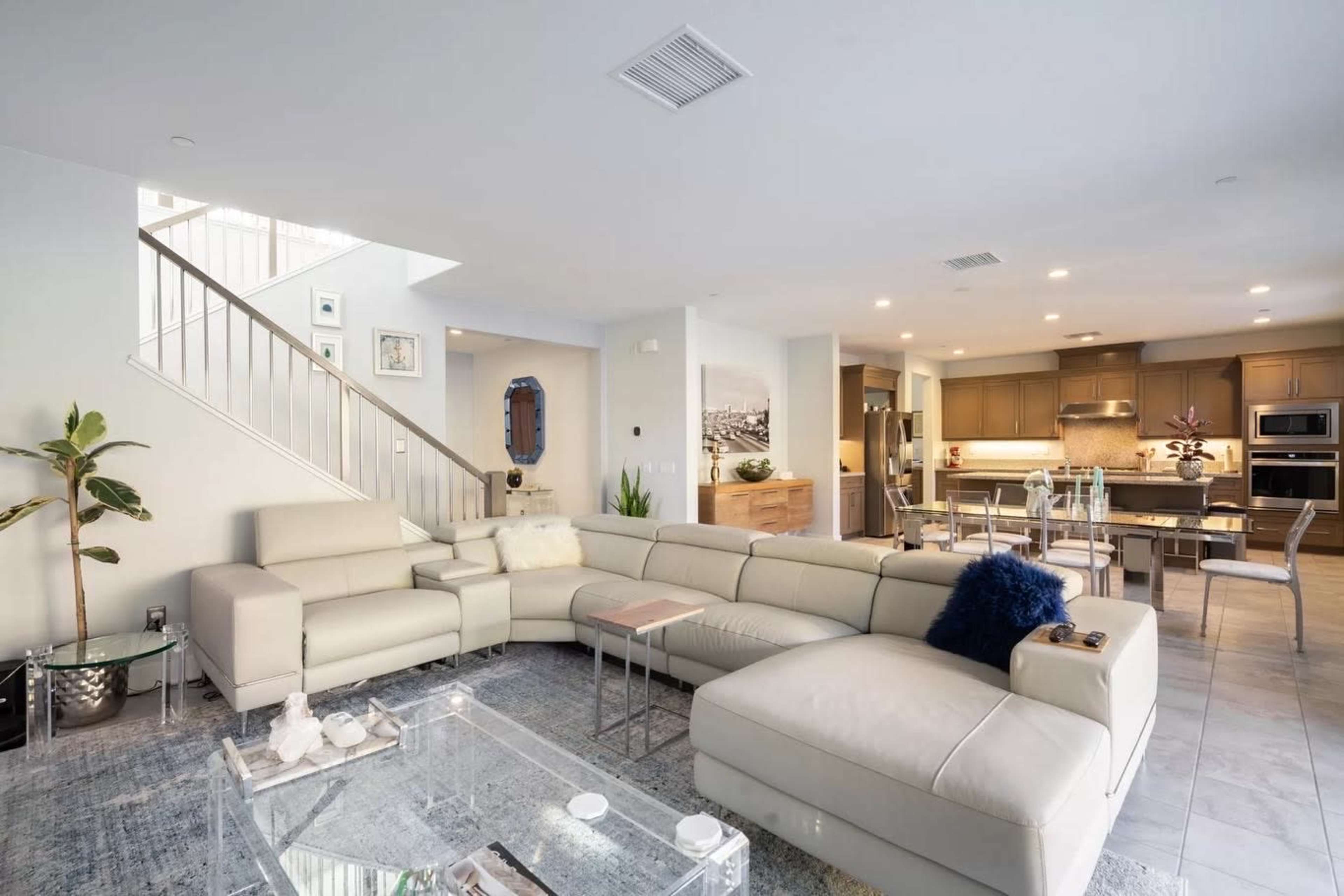 A modern living room features a large sectional sofa, a glass coffee table, and an open layout with kitchen access and a staircase.