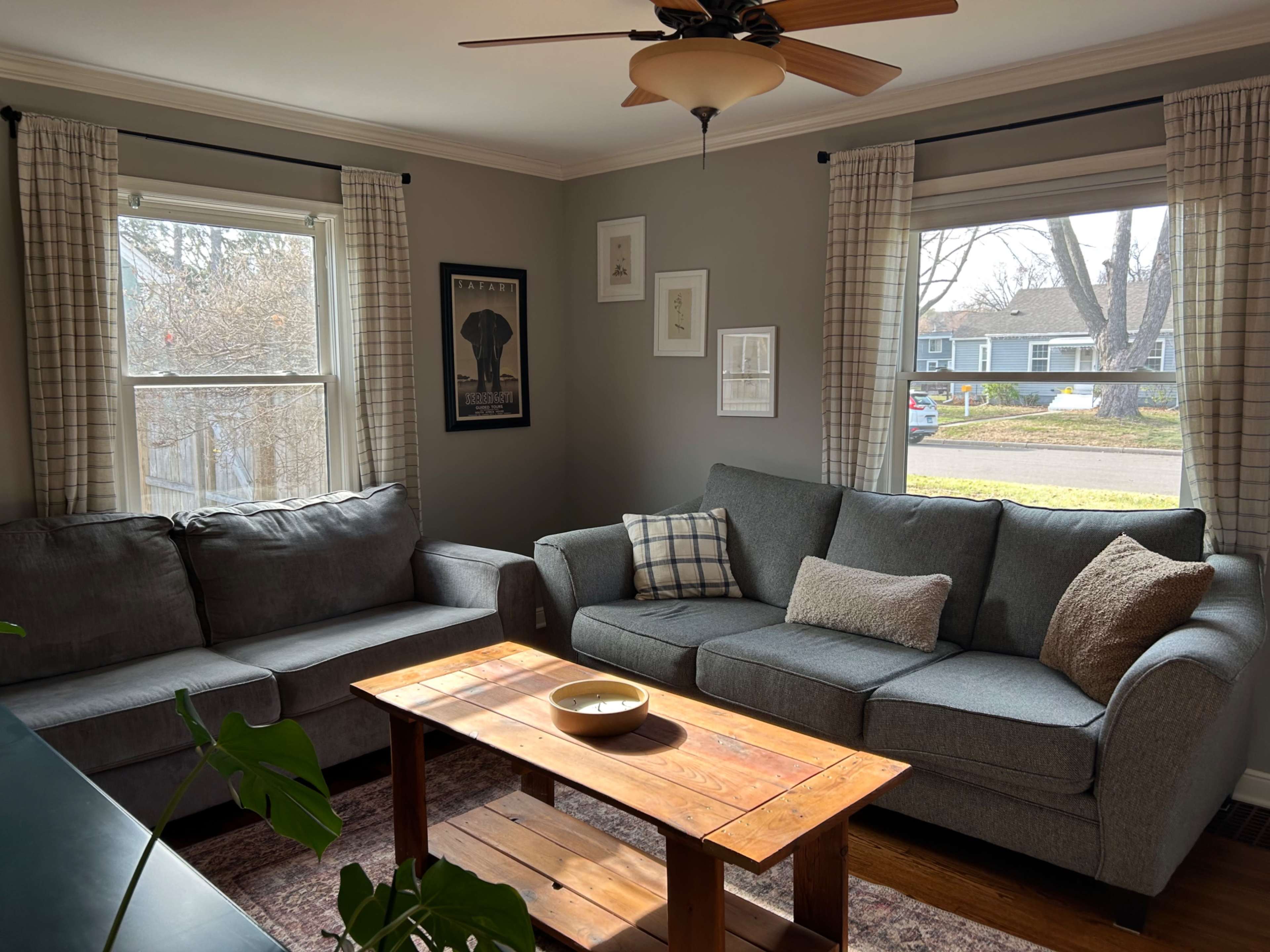 The image shows a cozy living room featuring a grey sectional couch, a wooden coffee table, and large windows with white curtains.