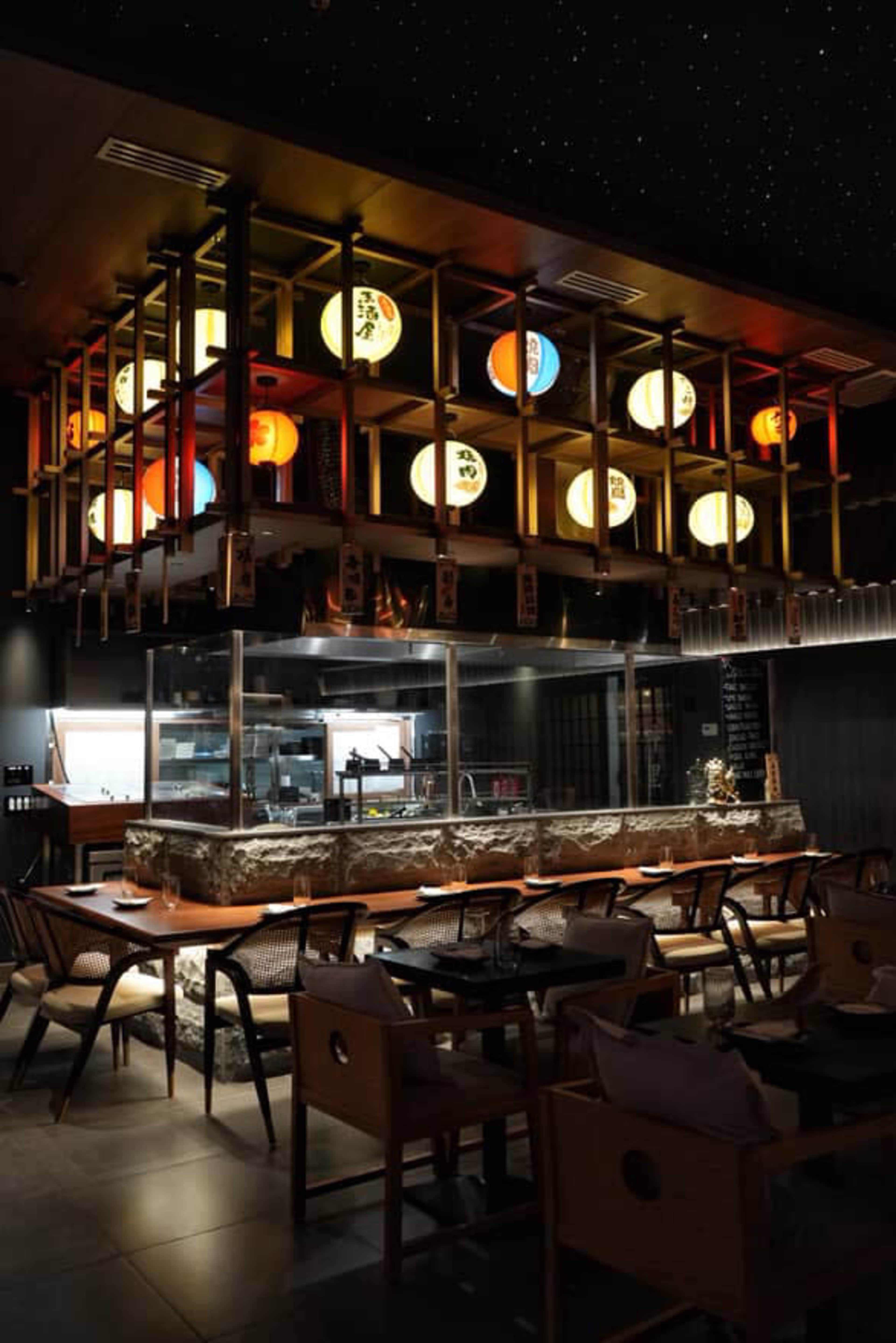 The image shows a modern restaurant interior with a prominently featured food preparation area, wooden seating, and decorative lanterns hanging above.