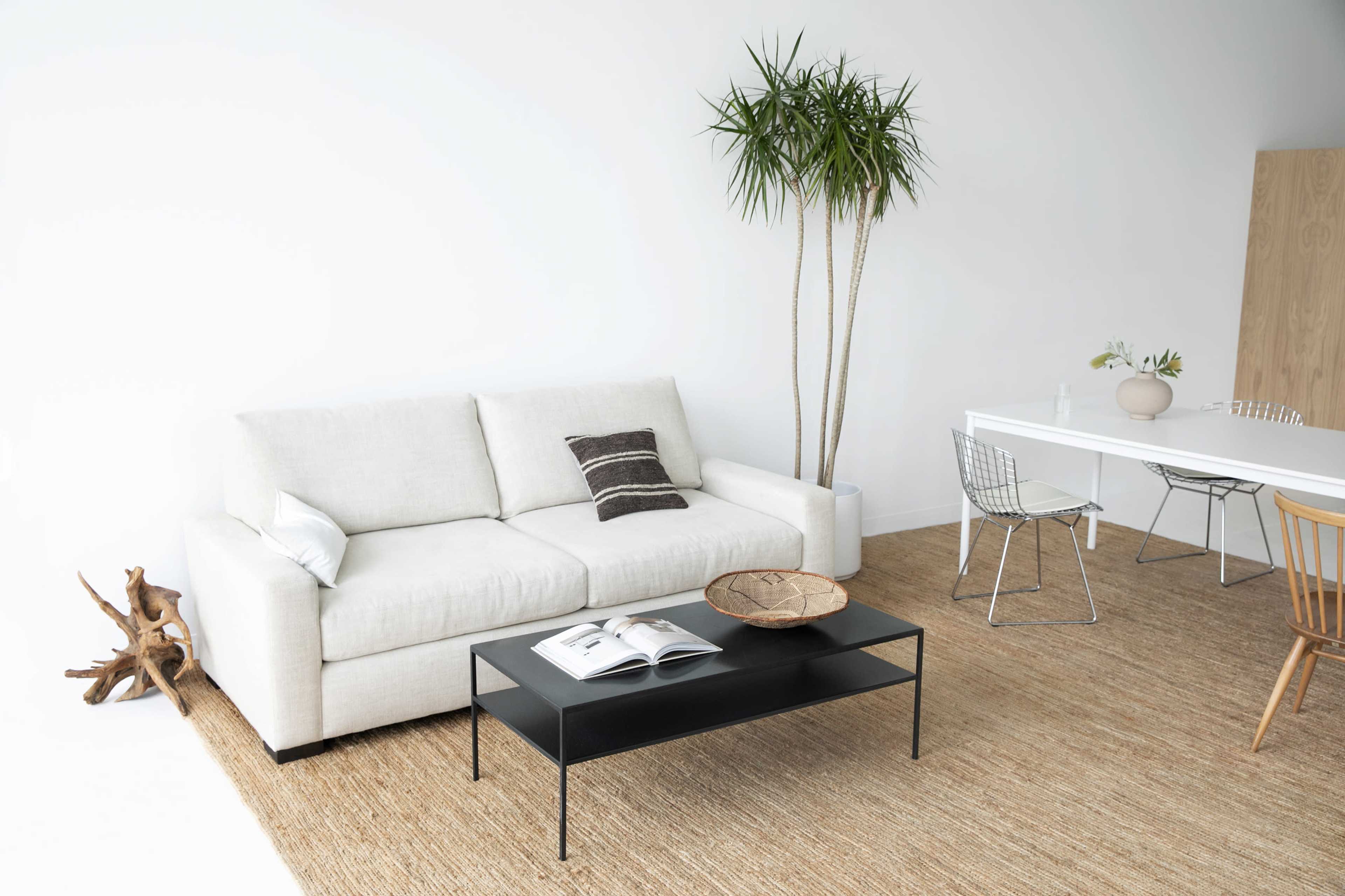 A light-colored sofa is positioned next to a black coffee table on a textured rug, with a dining table and chairs visible in the background.