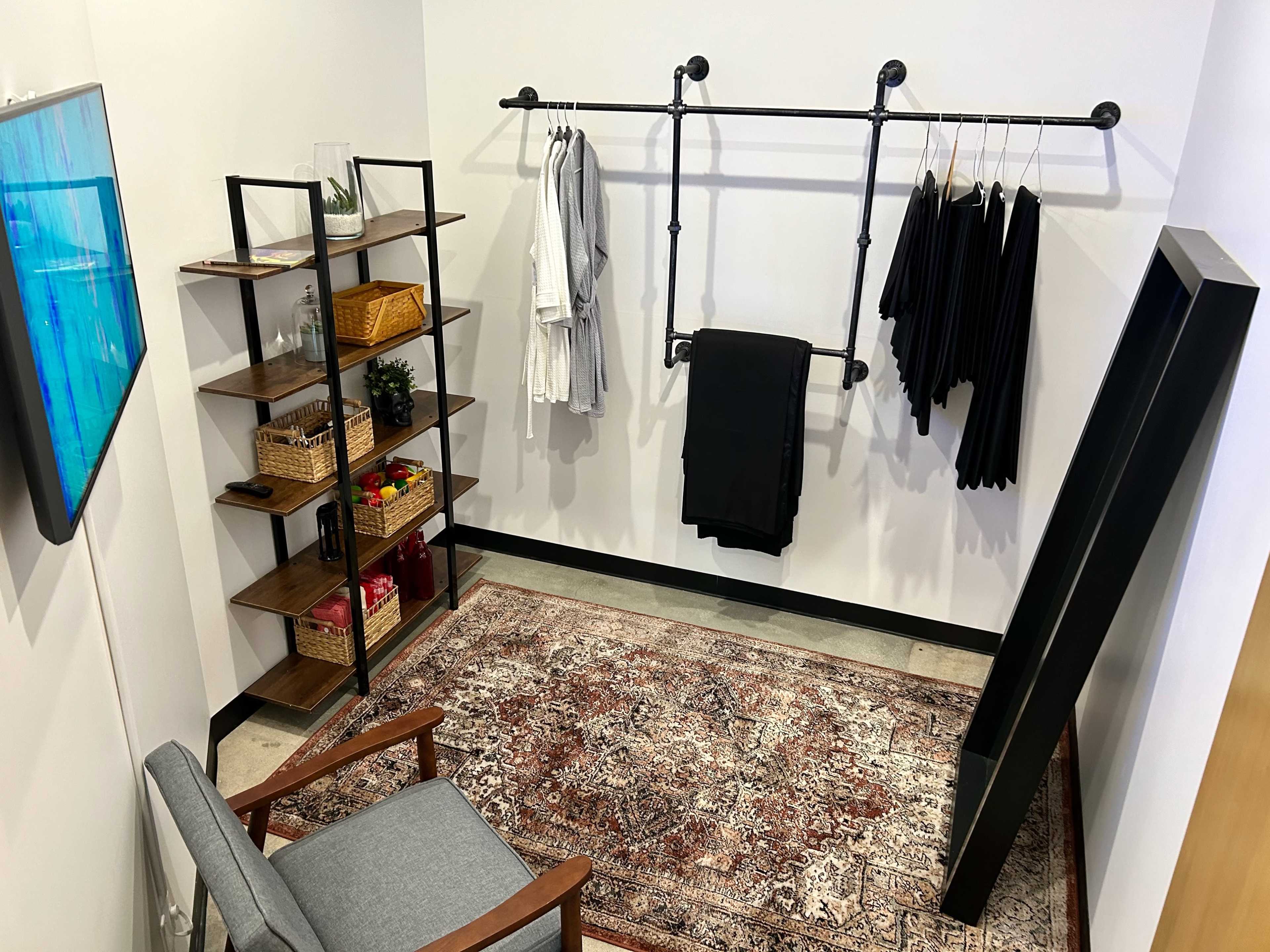A small, well-organized room featuring a clothing rack with hanging garments, a shelf with decorative baskets, a gray armchair, and a large wall-mounted TV.