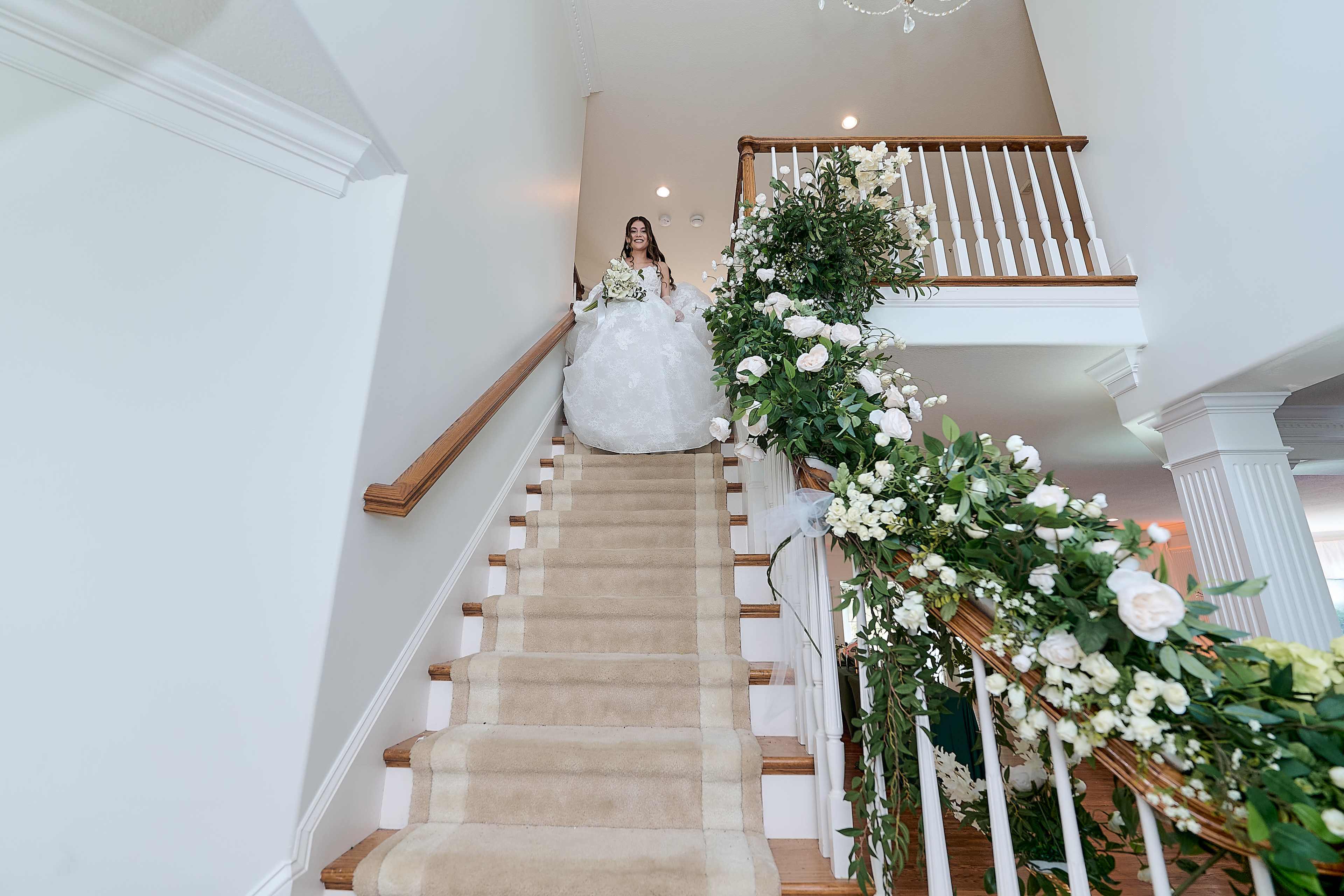 A bride in a white wedding dress stands at the top of a staircase adorned with flowers and greenery.