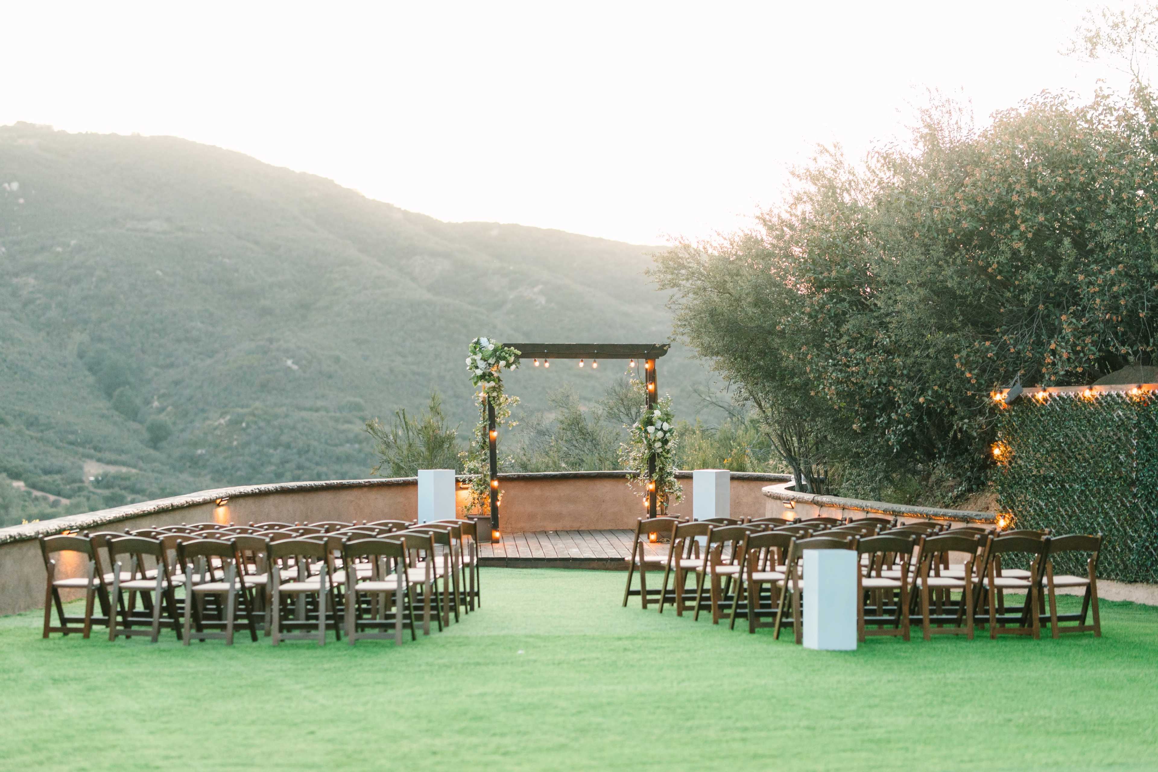 The image features an outdoor wedding ceremony setup with rows of wooden chairs facing a decorated arch against a scenic mountainous backdrop.