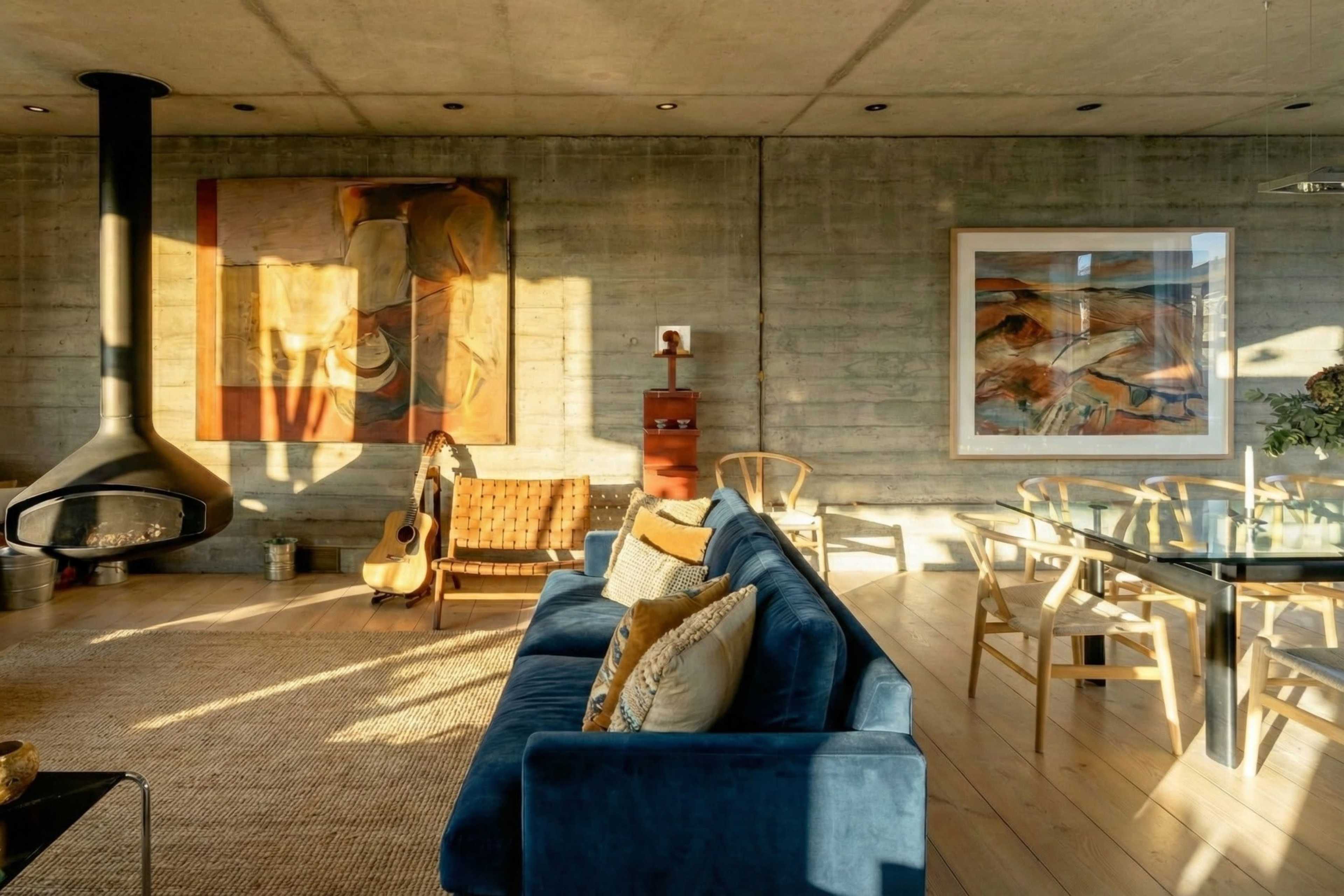 The image shows a modern living room featuring a concrete wall, a blue sofa, a glass dining table, and artwork on the walls.
