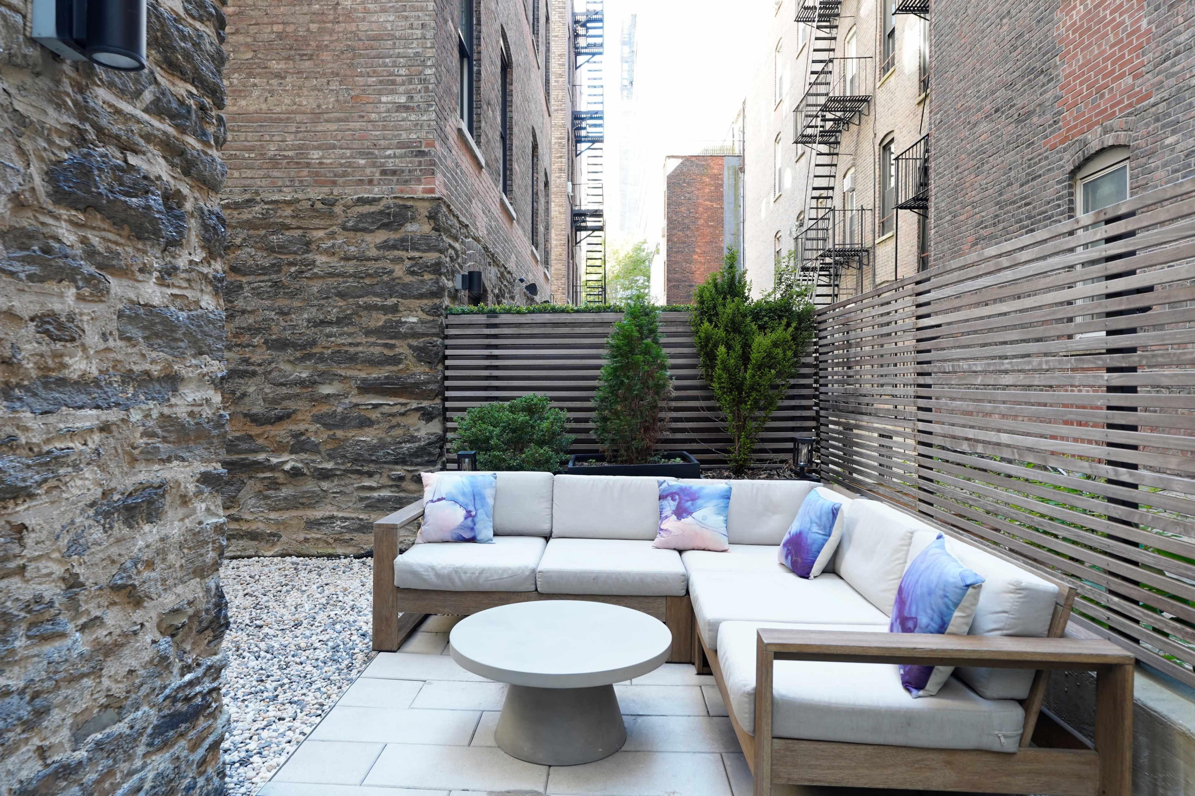 The image features a small outdoor patio with a sectional sofa, a round table, and greenery, enclosed by brick and wooden privacy screens.