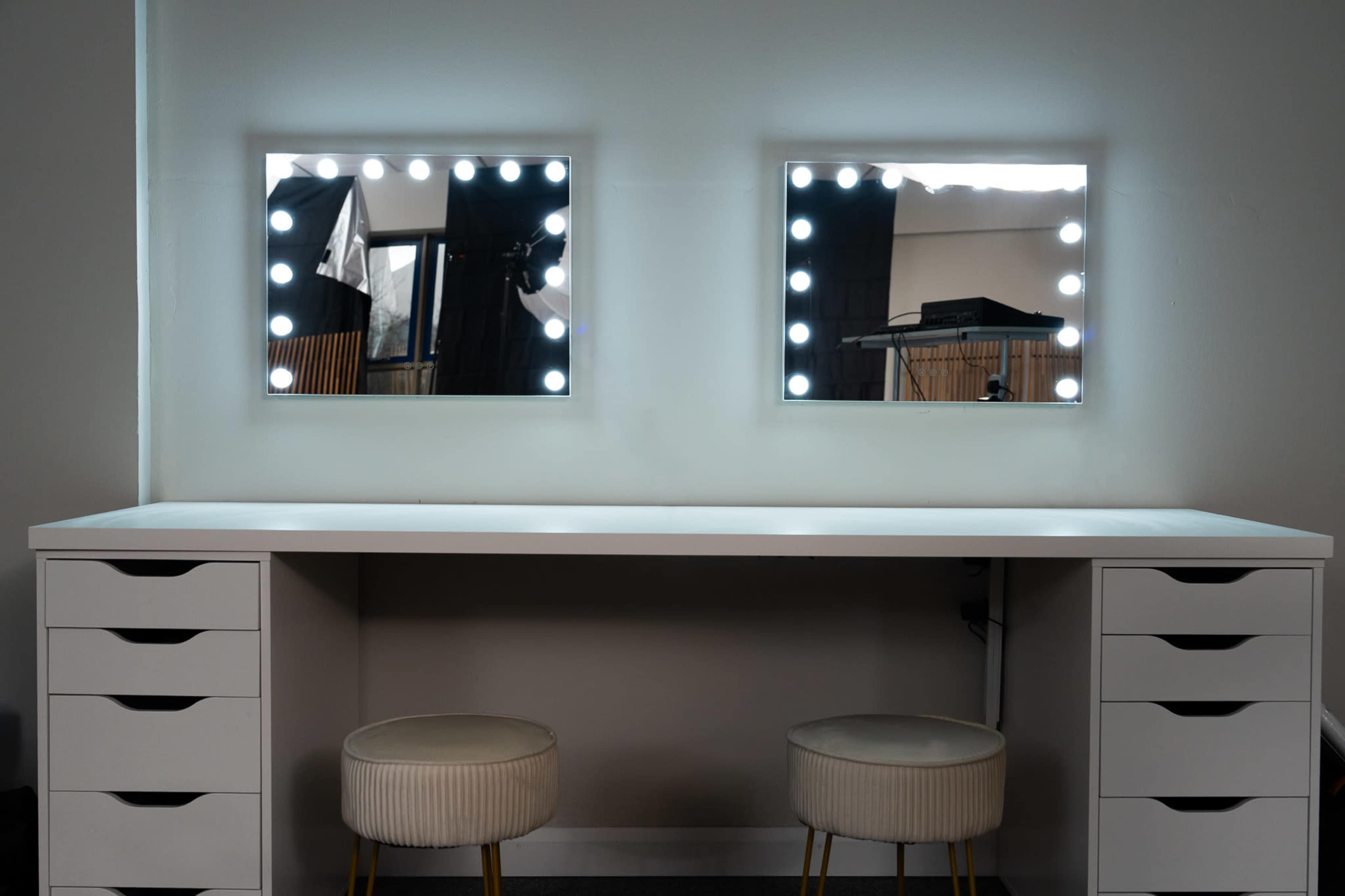 The image shows a minimalist white vanity desk with two illuminated mirrors and two stools in front.