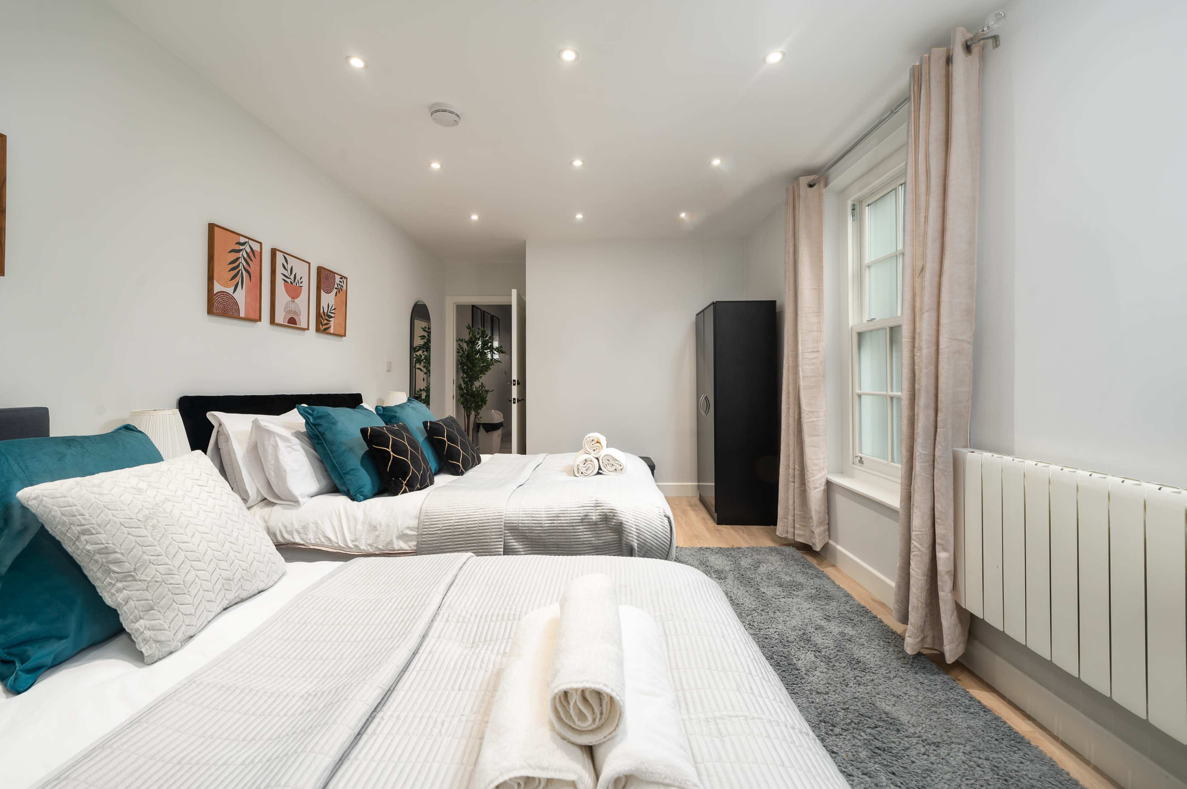 The image shows a well-lit bedroom featuring two beds with neatly arranged linens, a black wardrobe, and potted plants.