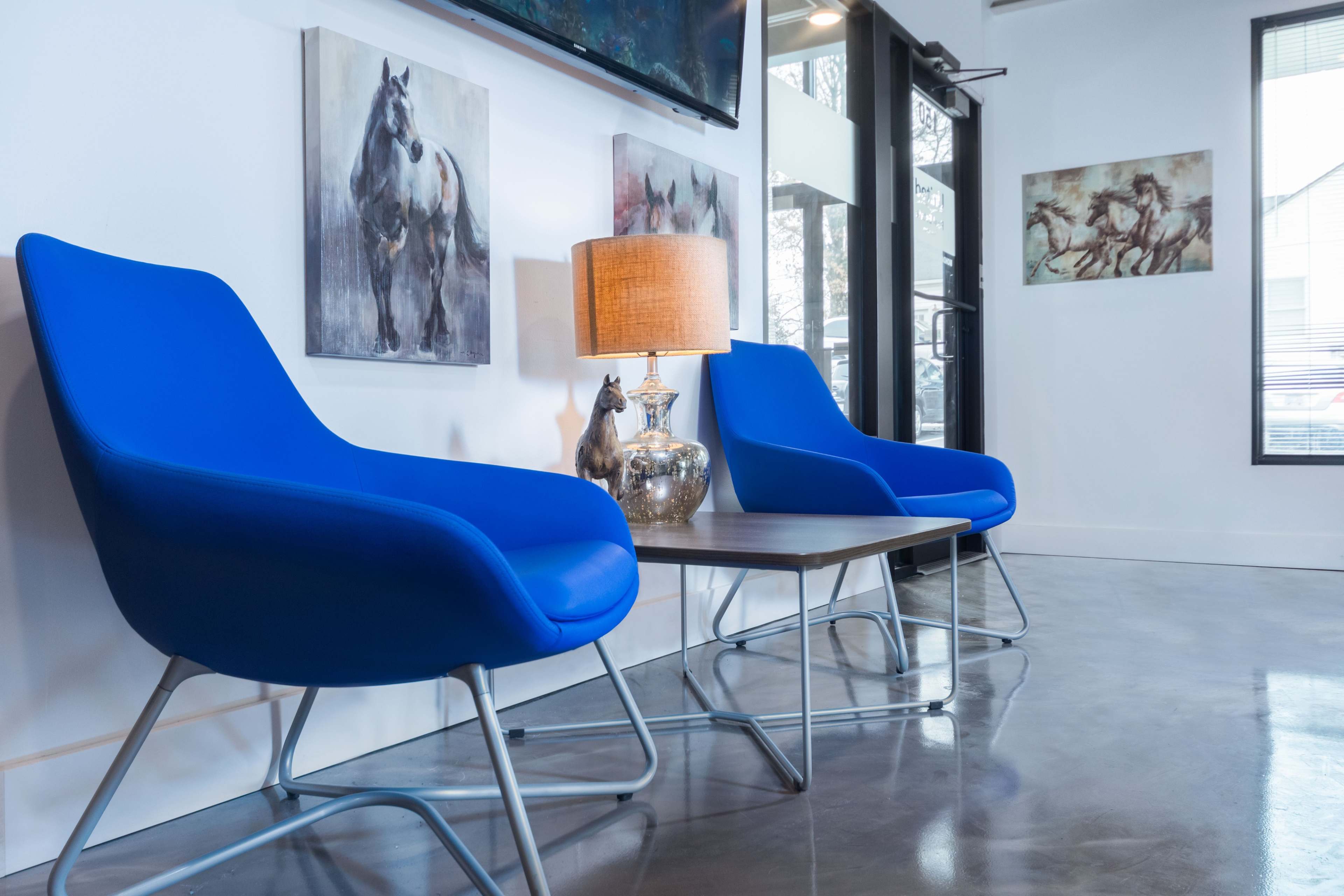 Two blue chairs are positioned beside a wooden table, with a lamp and decorative items, in a brightly lit modern room featuring horse-themed artwork on the walls.