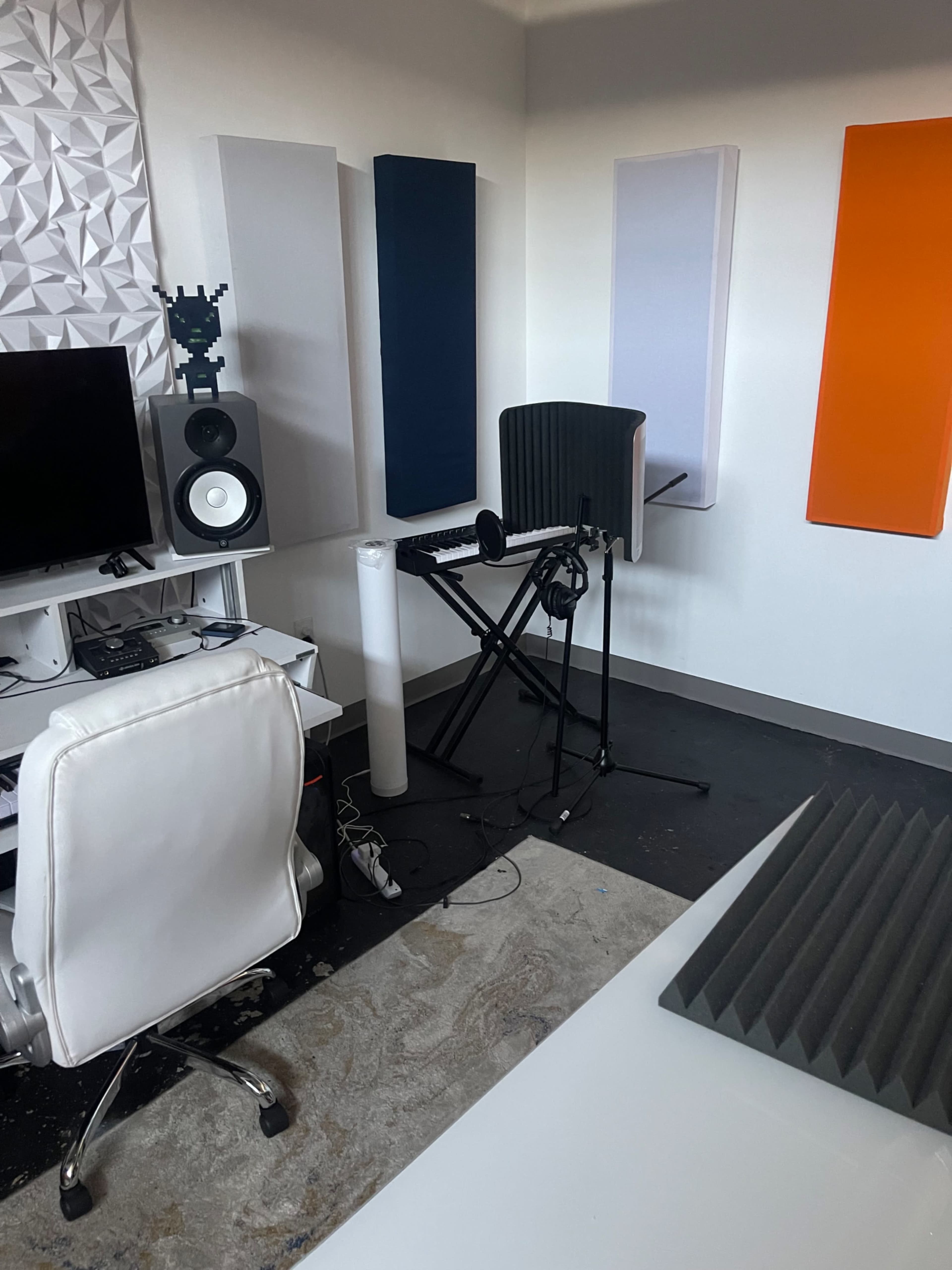 The image shows a music production workspace featuring soundproofing panels on the walls, a microphone stand, a keyboard, and a computer setup.