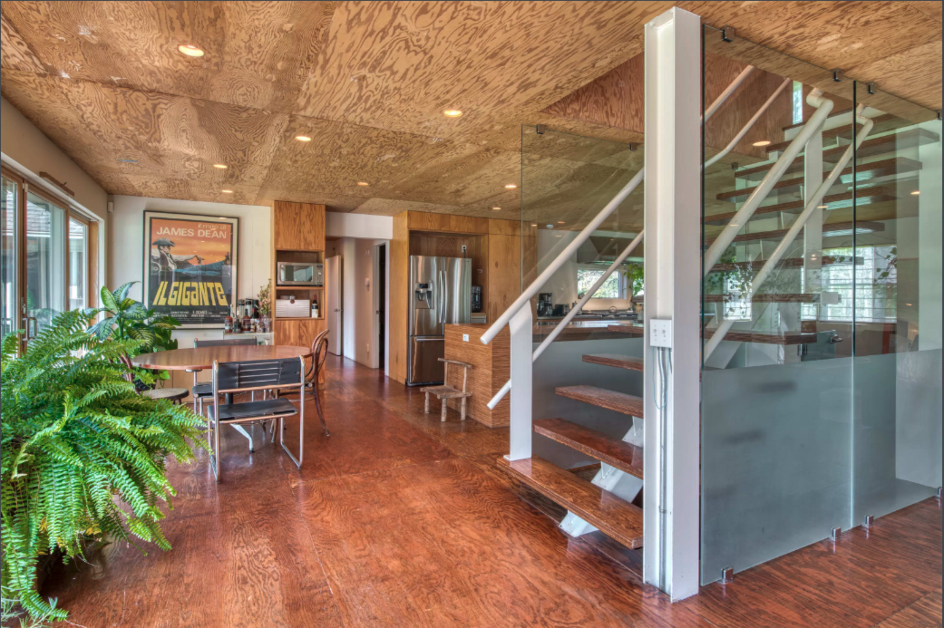 A modern interior space featuring a dining area, kitchen, and a staircase with glass railing, all complemented by wood flooring and ceiling.