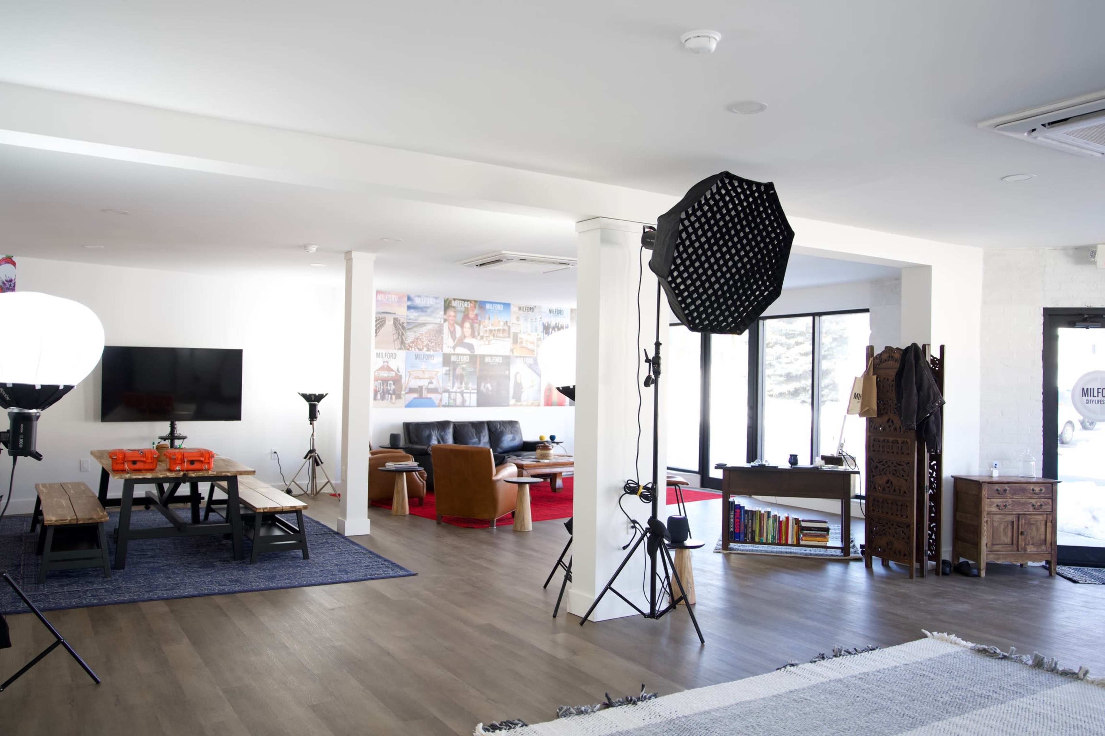 The image shows a spacious, modern studio or living area featuring various seating arrangements, a large wall-mounted TV, and professional lighting equipment set up for a shoot.