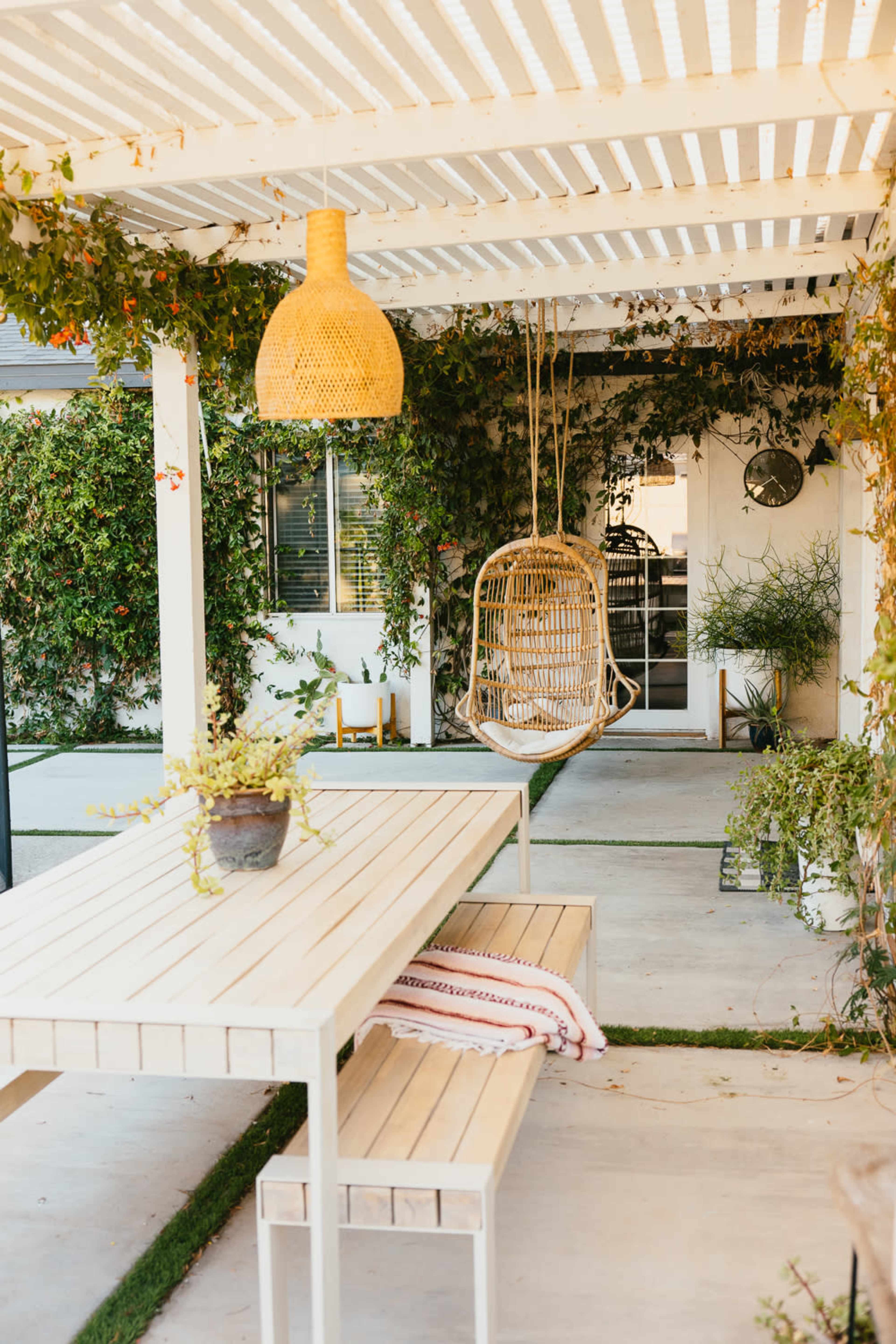 A covered outdoor patio features a wooden table and benches, a hanging wicker chair, and lush greenery along the walls.
