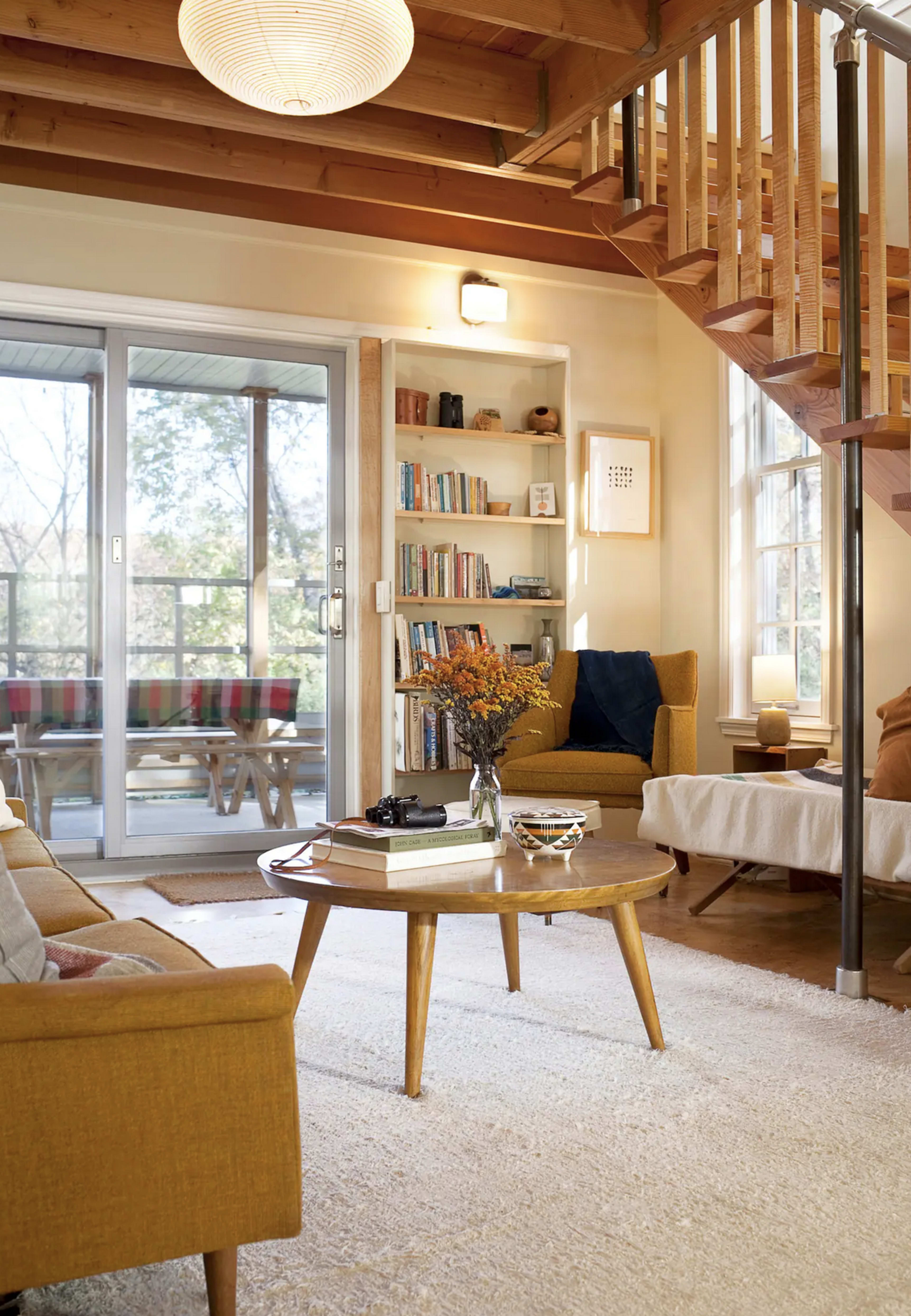 The scene features a cozy living room with a staircase, a wooden coffee table, and bookshelves filled with books, illuminated by natural light from large windows.