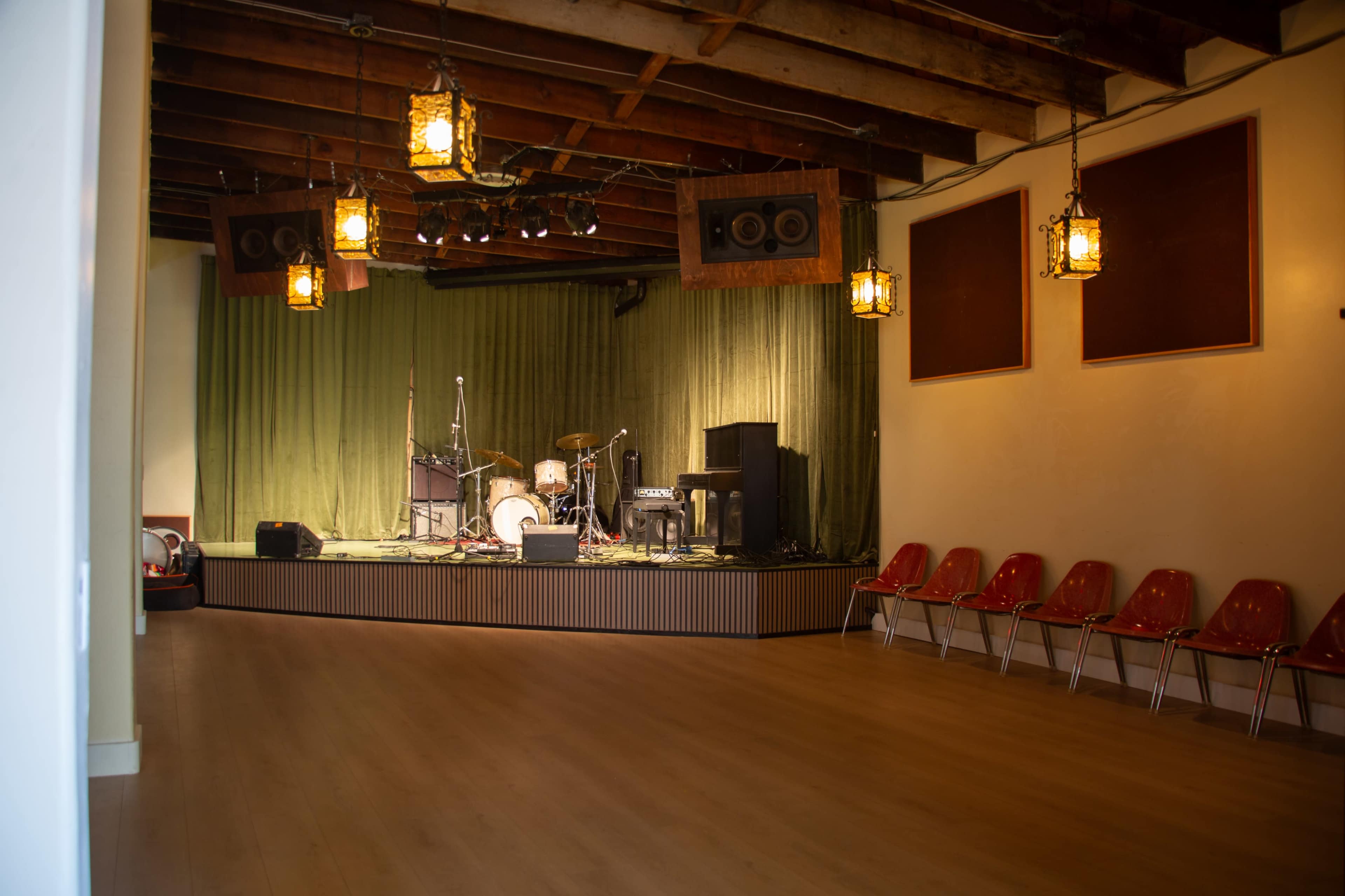 The image shows a stage set up for a performance, featuring musical instruments, a green backdrop, and several empty red chairs arranged in front of it.