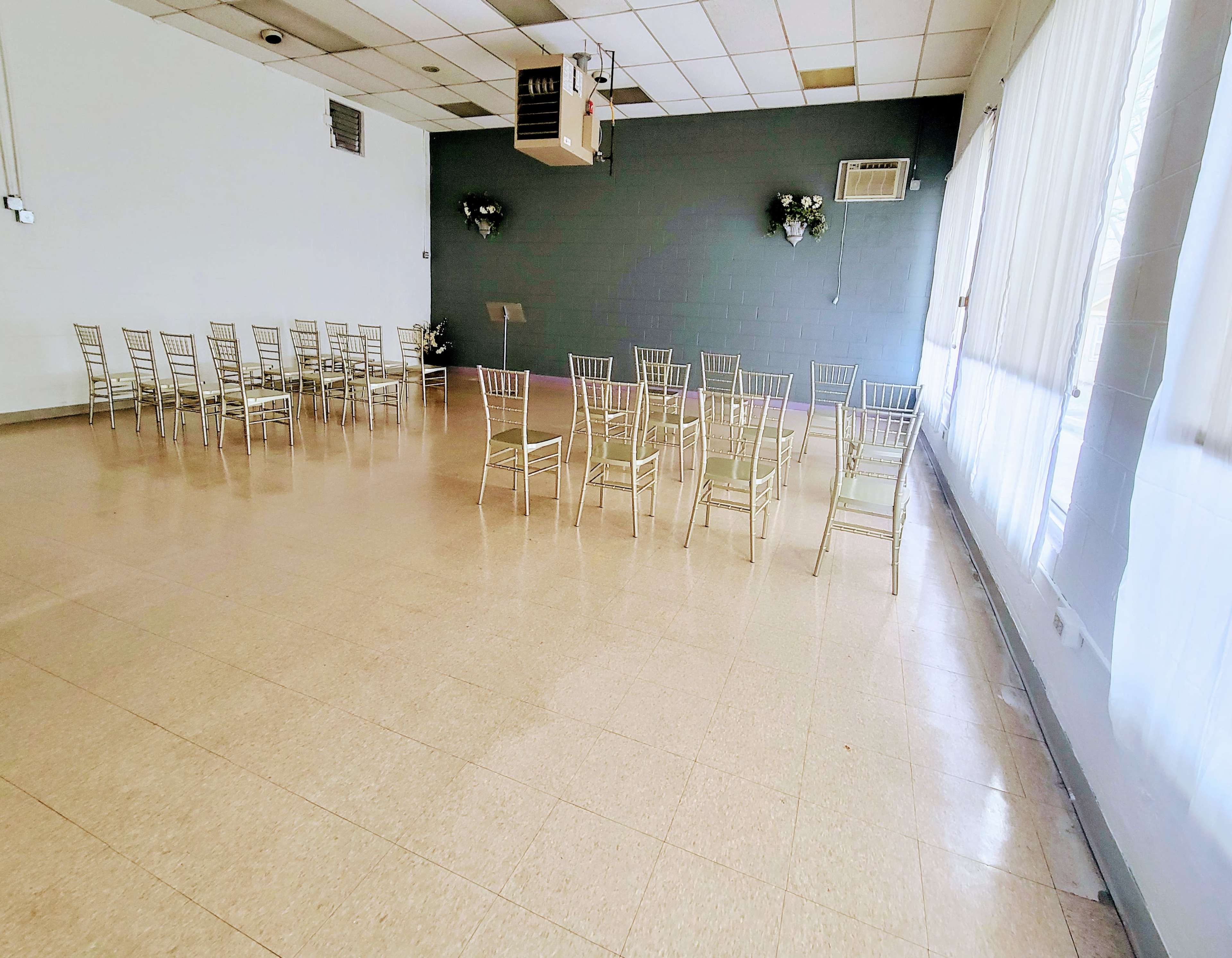 A spacious, empty room with rows of metal chairs arranged facing a blank wall, large windows allowing natural light to enter.