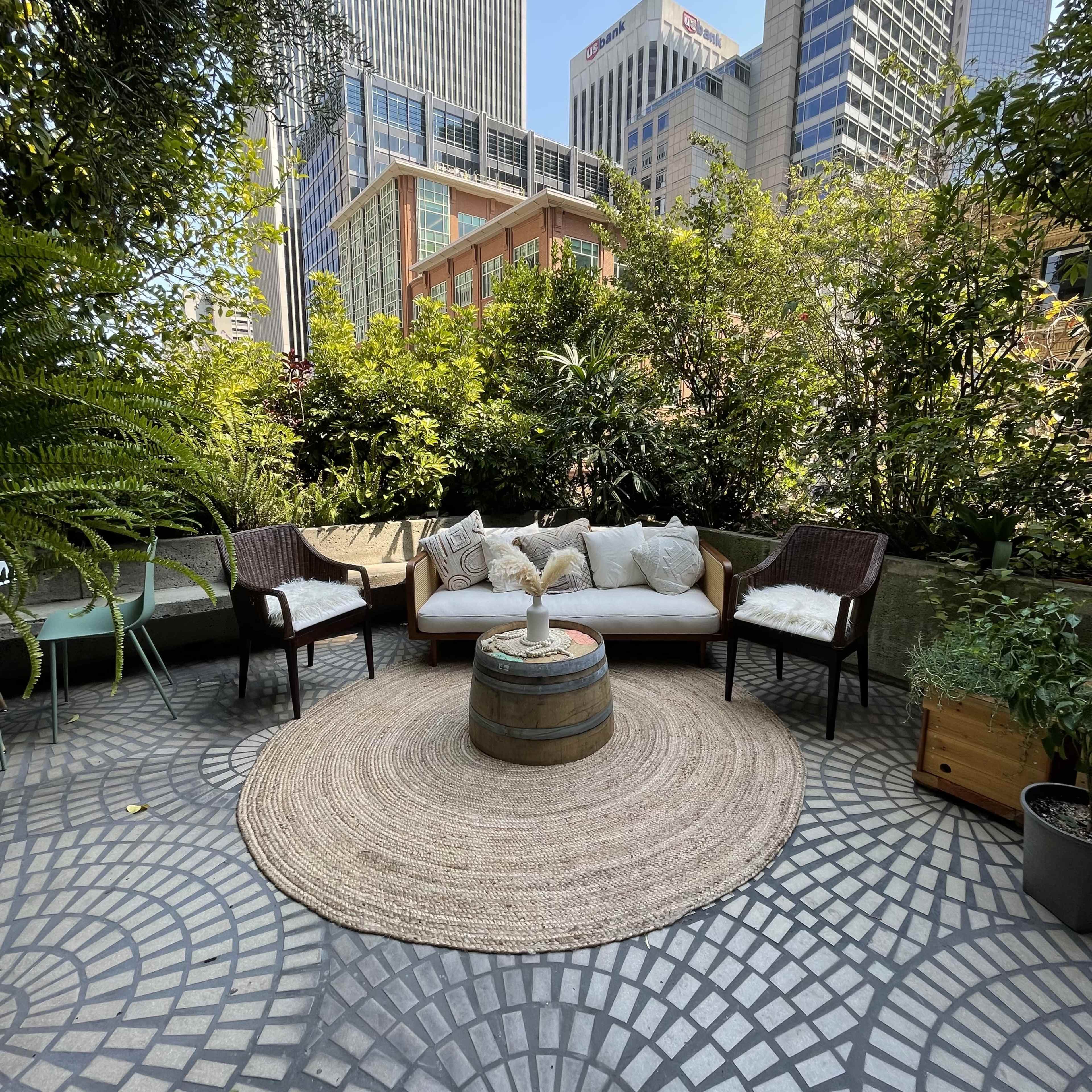 A cozy outdoor seating area features a circular rug, a barrel table, a sofa with cushions, and two chairs surrounded by greenery and city buildings in the background.