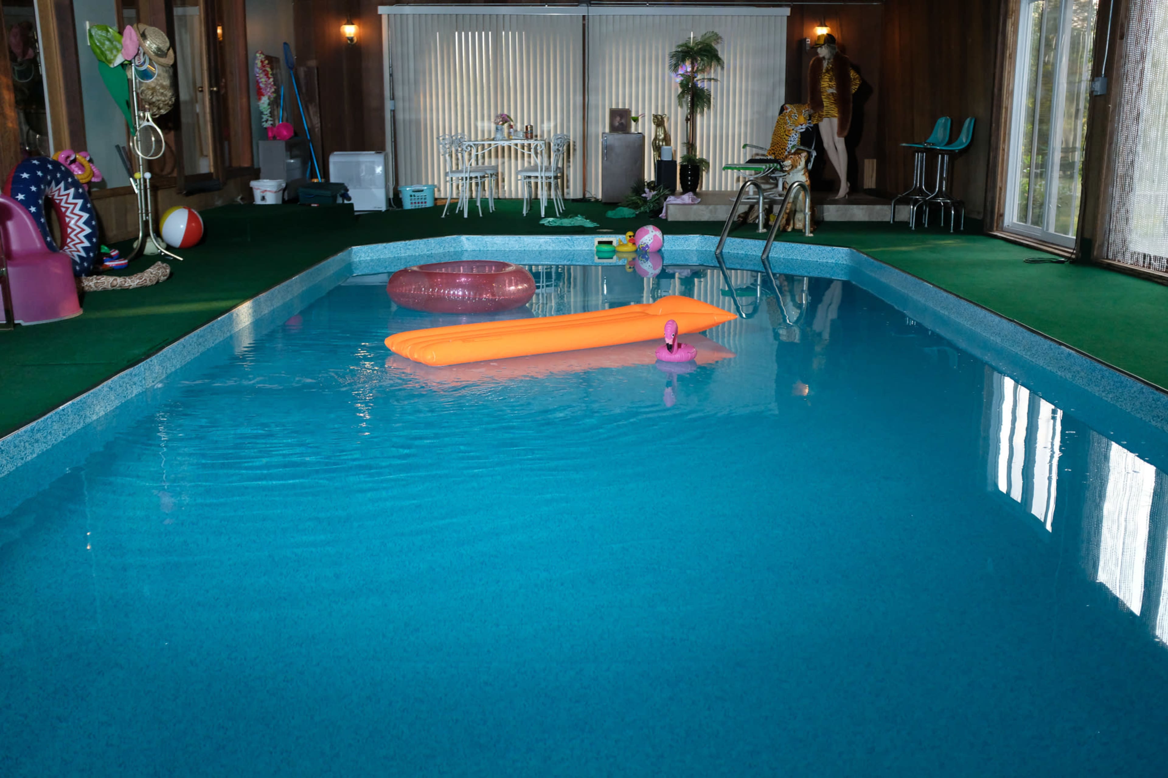 The image shows an indoor pool surrounded by green carpet, with a pink flamingo float and an orange float in the water, and a decorative area featuring furniture and plants in the background.