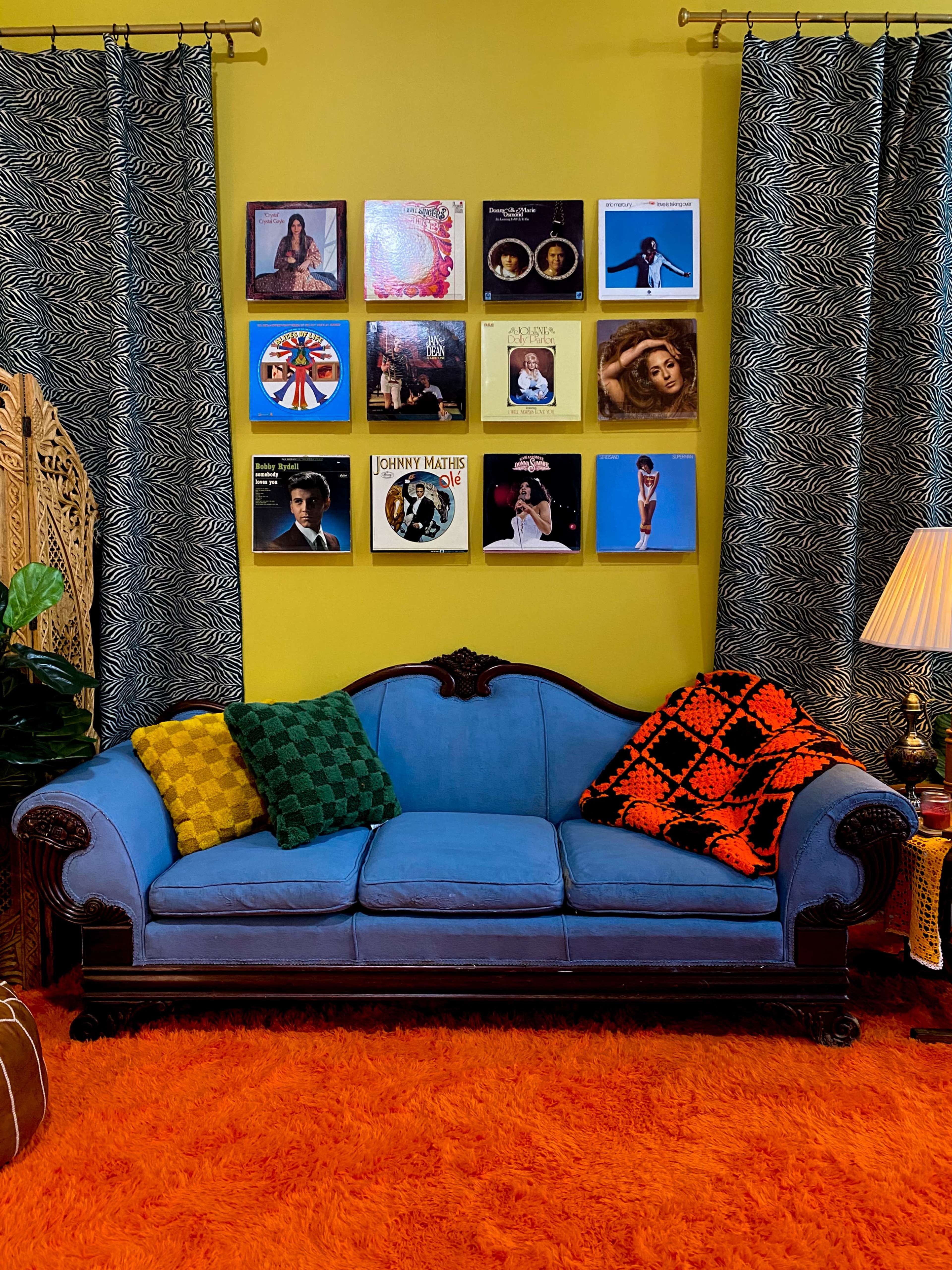 A blue vintage couch is centered in front of a wall adorned with framed album covers, while orange shag carpet and patterned curtains complement the scene.