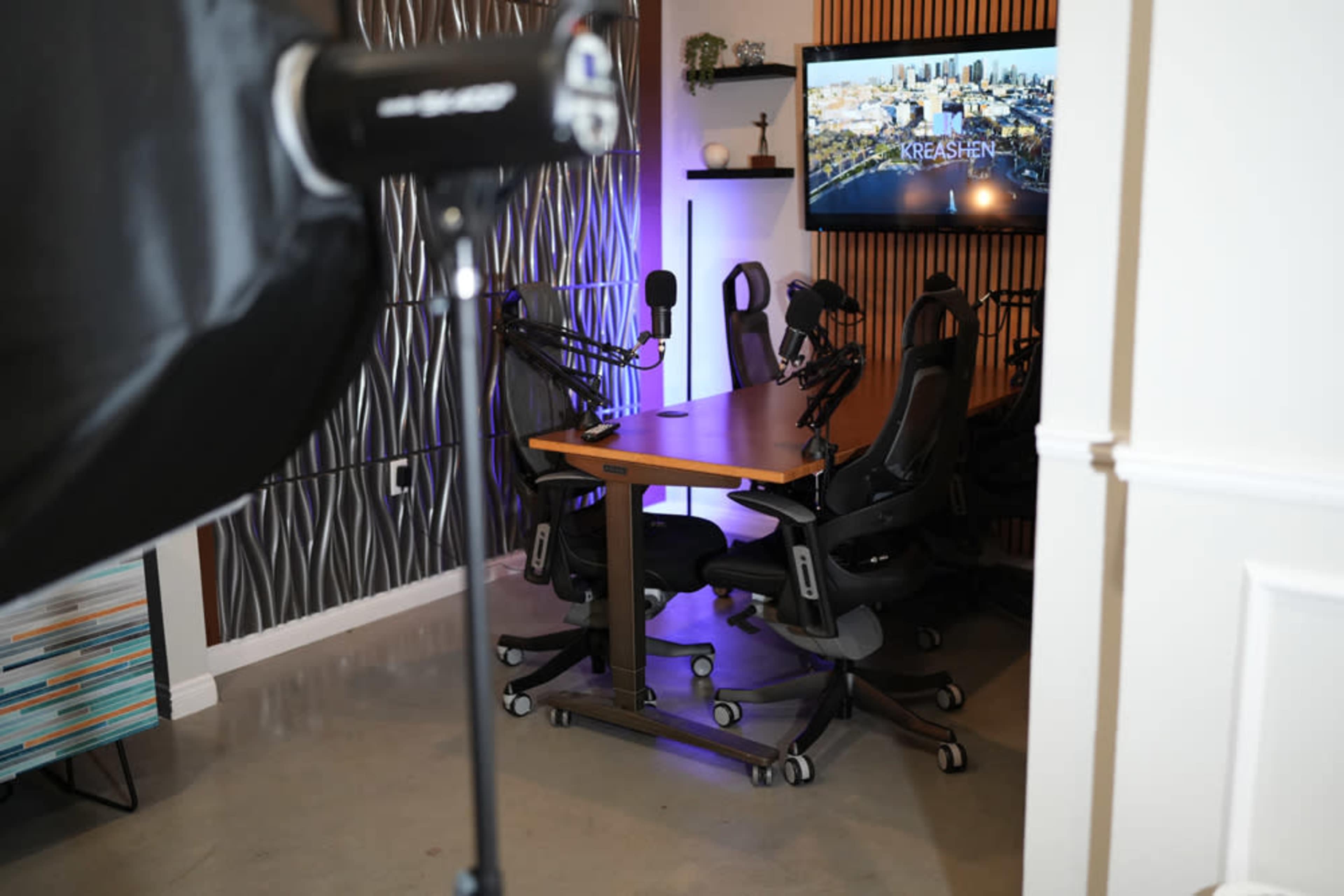 The image shows a podcast studio featuring a wooden table surrounded by black chairs and equipped with multiple microphones and a large screen displaying a cityscape.