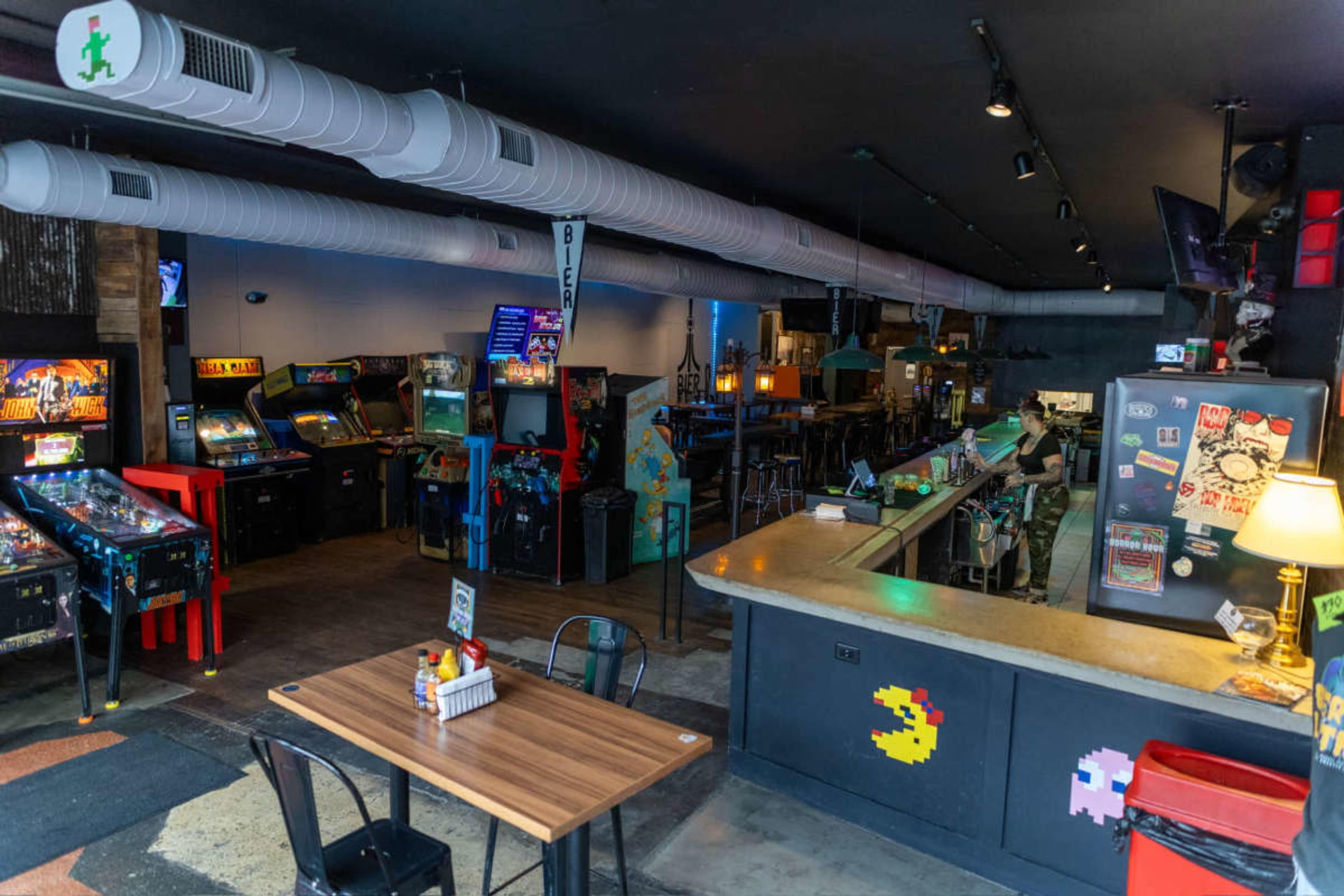 The image shows a retro-style arcade bar with arcade machines on one side, a bar counter, and tables for seating.