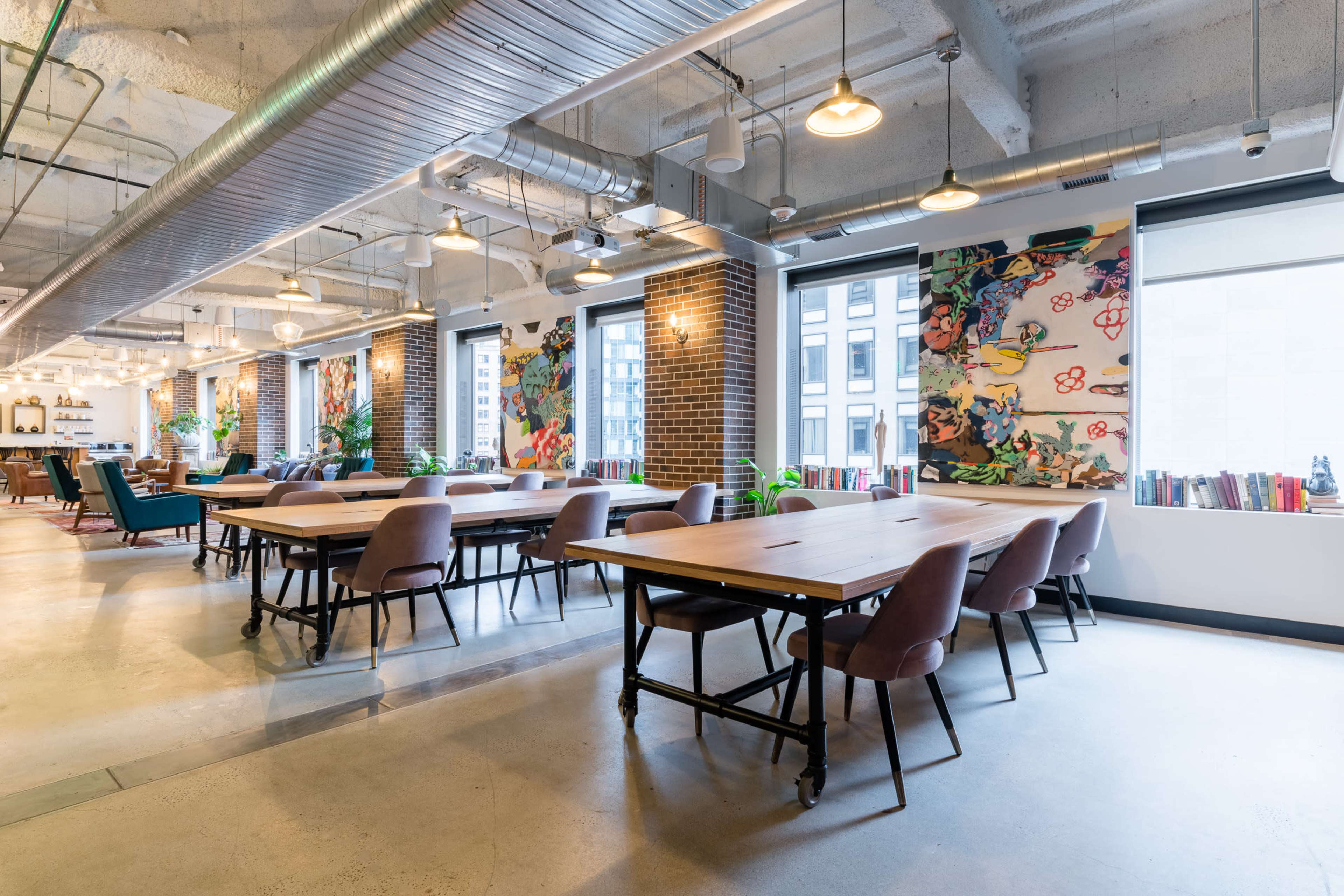 The image shows a modern open office space featuring wooden tables, pink chairs, and large windows with vibrant artwork on the walls.