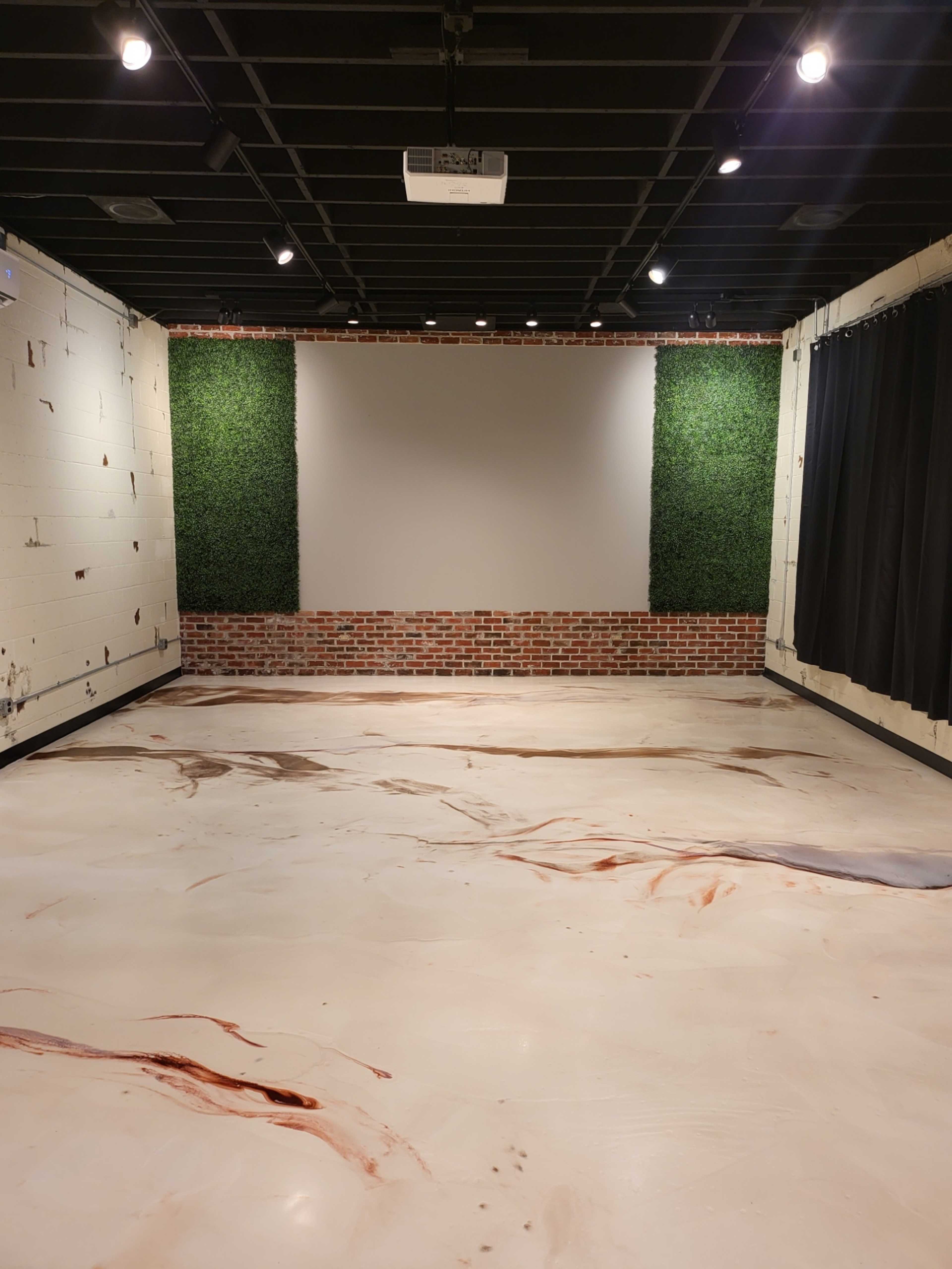 A spacious room with a smooth, light-colored floor featuring streaks, a brick wall with greenery on either side, and a blank wall at the front used for presentations.