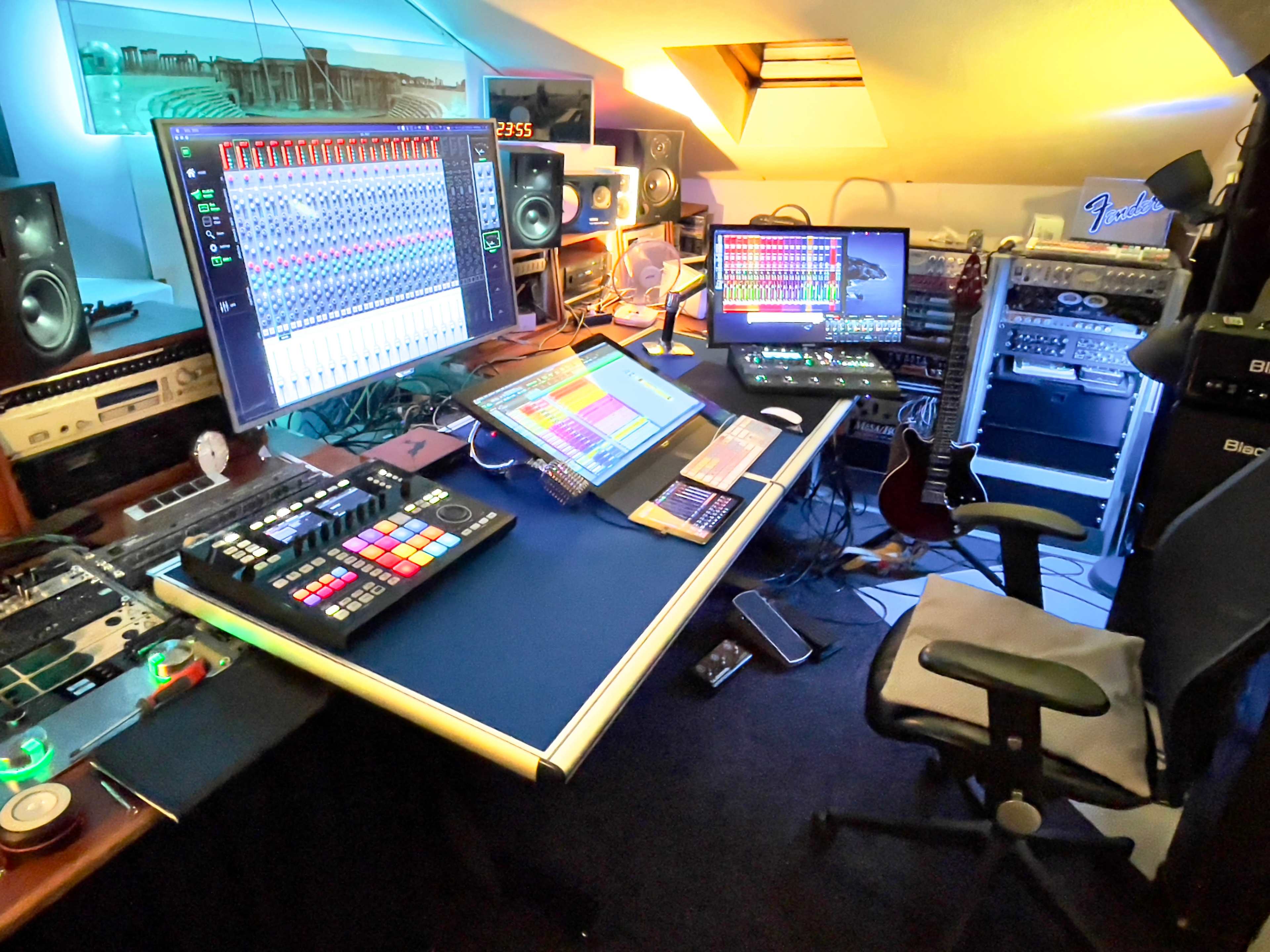 A modern home music studio with multiple screens, audio equipment, and musical instruments arranged on a desk.