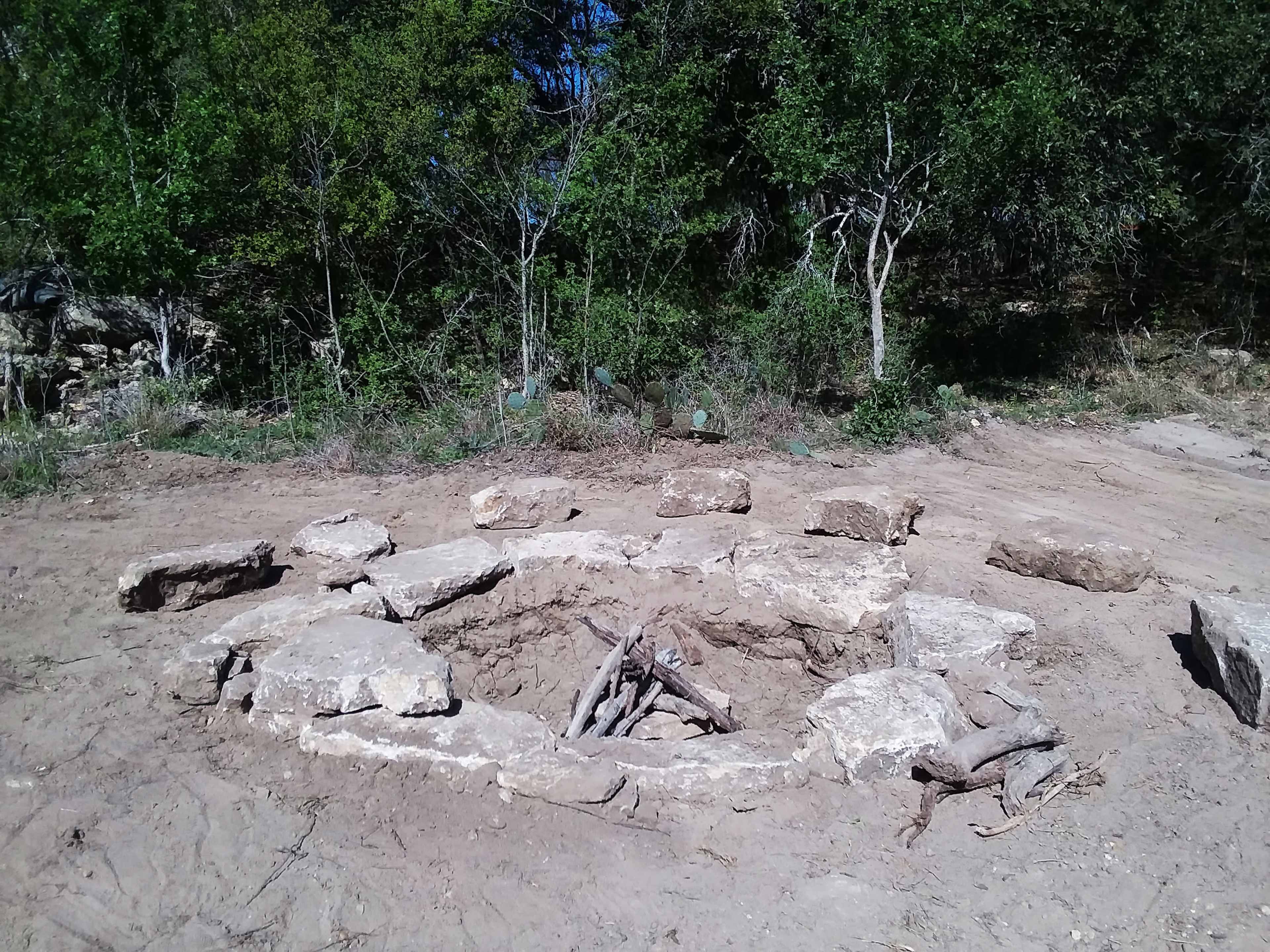 The image shows a circular fire pit made of large stone slabs, surrounded by a dirt area with sparse vegetation.