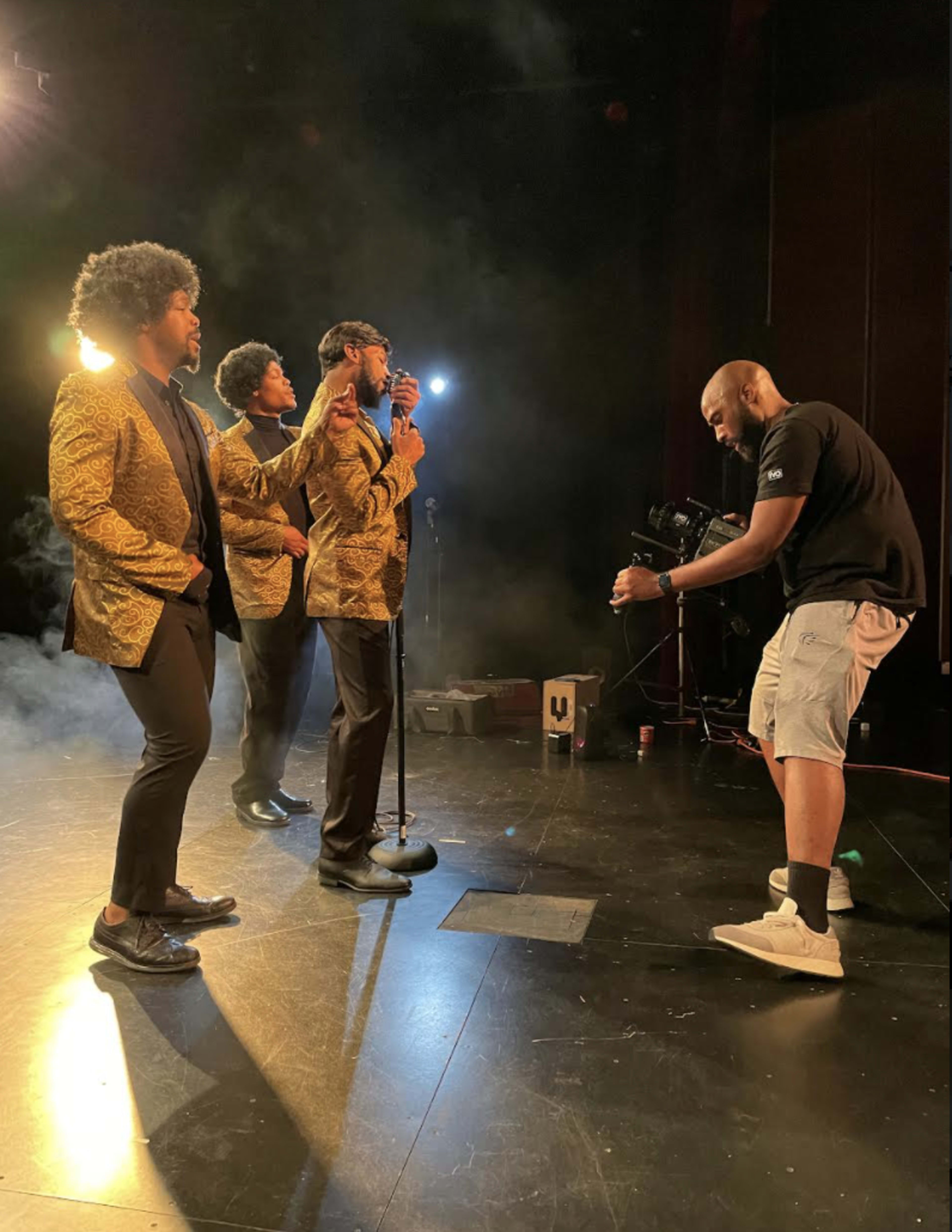 Three singers in golden jackets perform on stage while a crew member operates a camera.