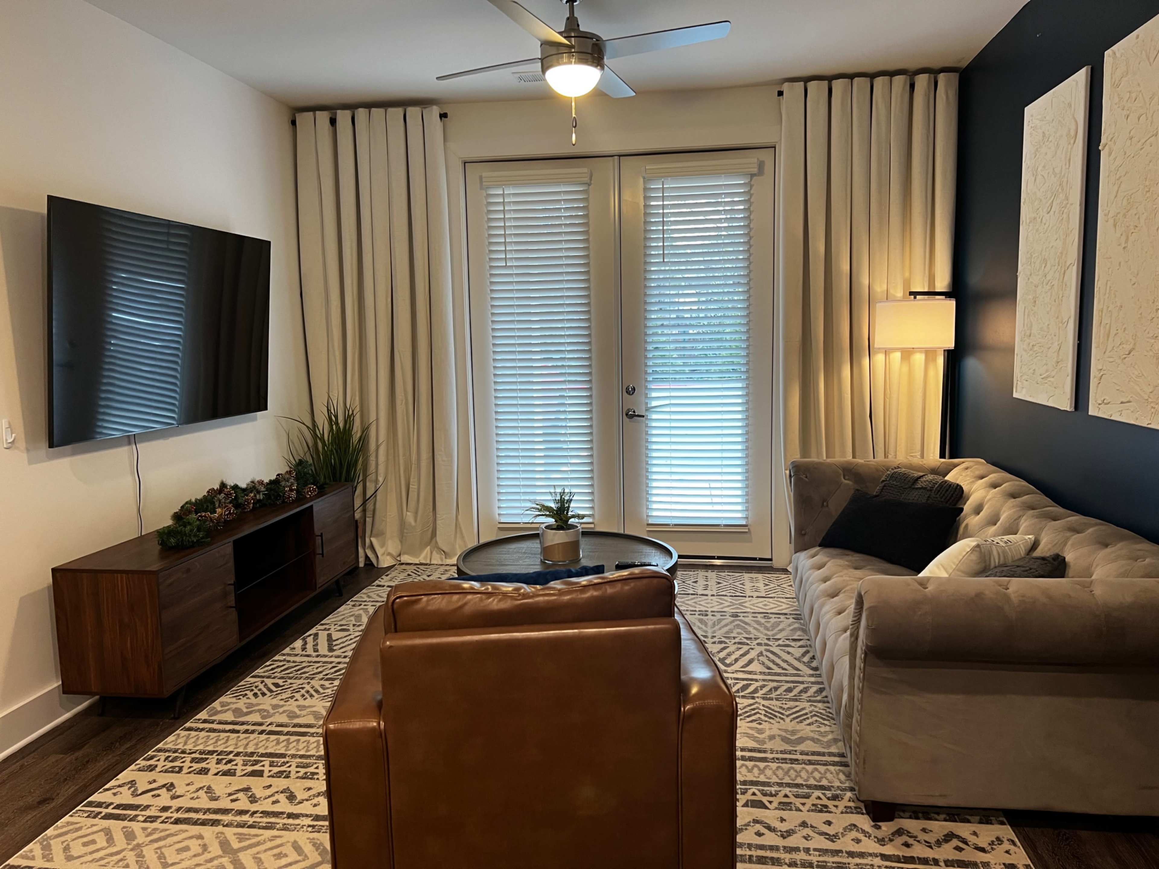 The living room features a television mounted on the wall, a beige sofa, a brown armchair, and large windows covered with sheer curtains.