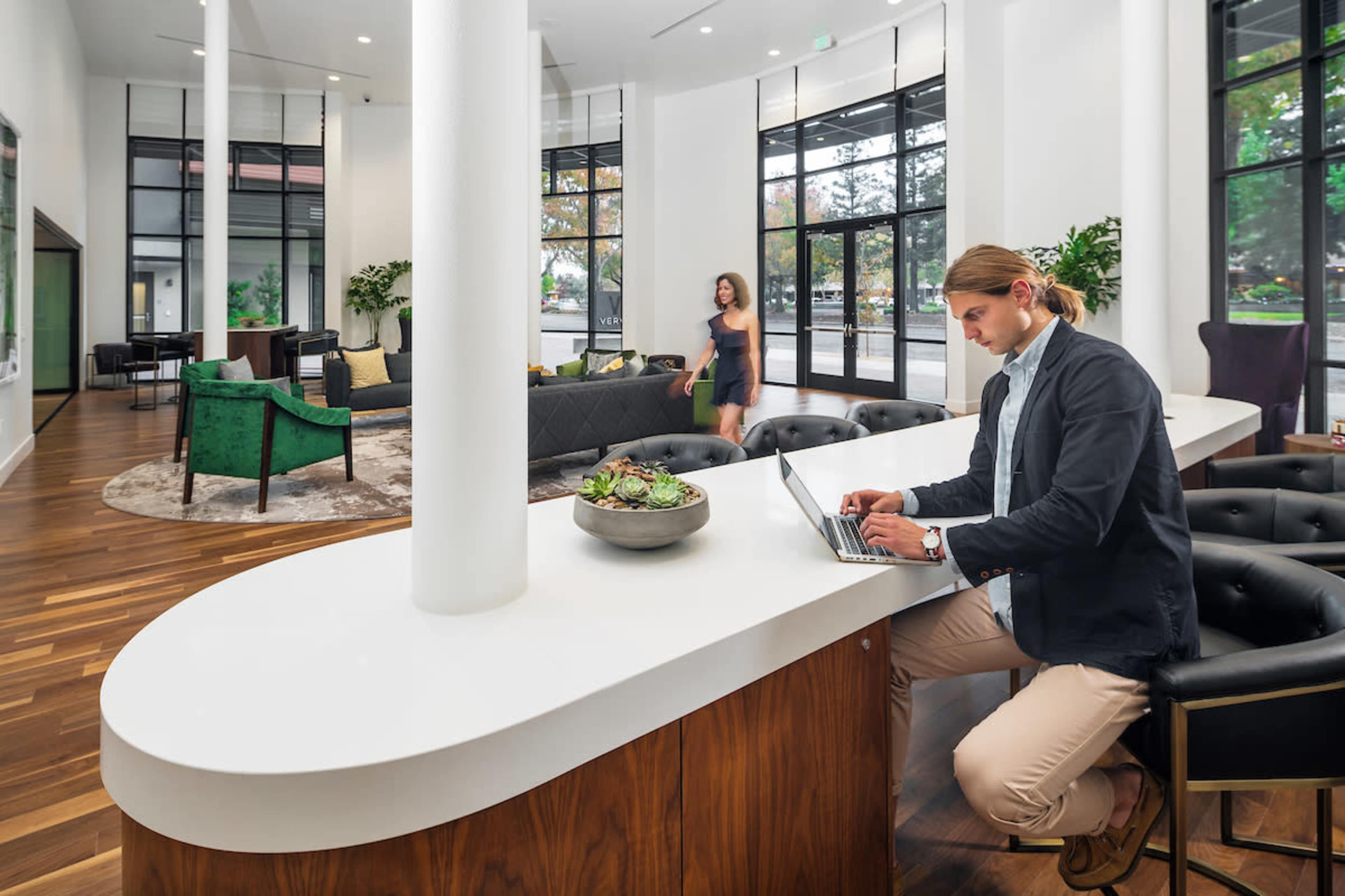 A man works on a laptop at a curved white countertop while a woman walks through a modern lobby with large windows and stylish seating.