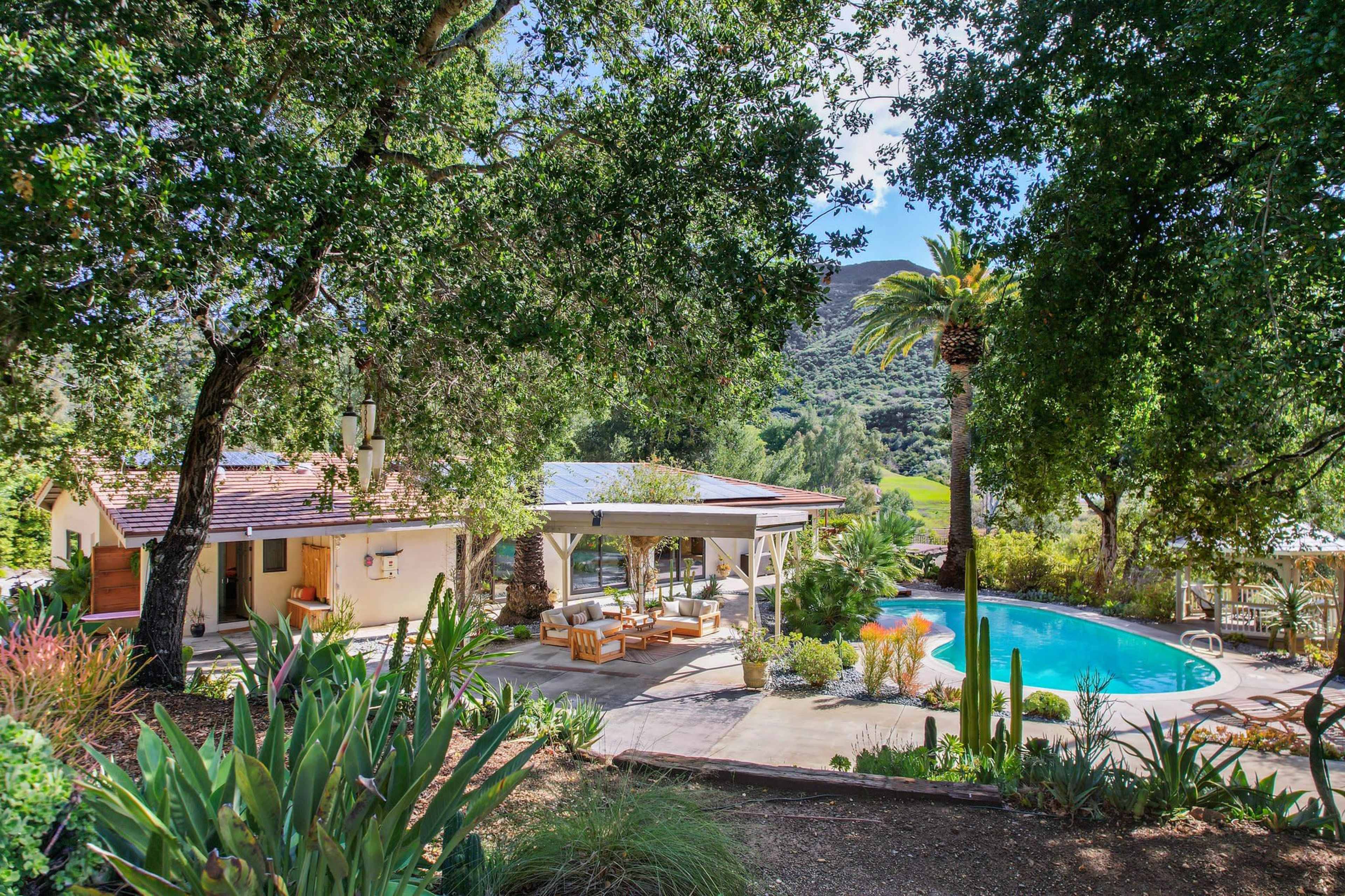 A landscaped outdoor area features a swimming pool surrounded by seating and lush greenery, with a house and a large tree in the background.