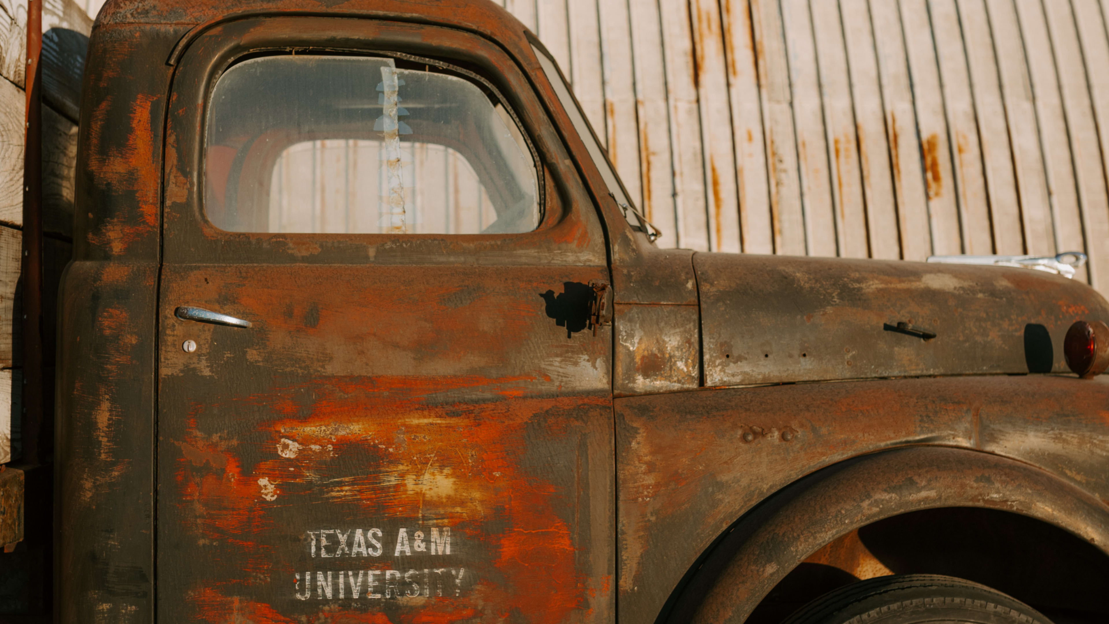 An old, rusted truck with a faded Texas A&M University logo is parked beside a wooden fence.