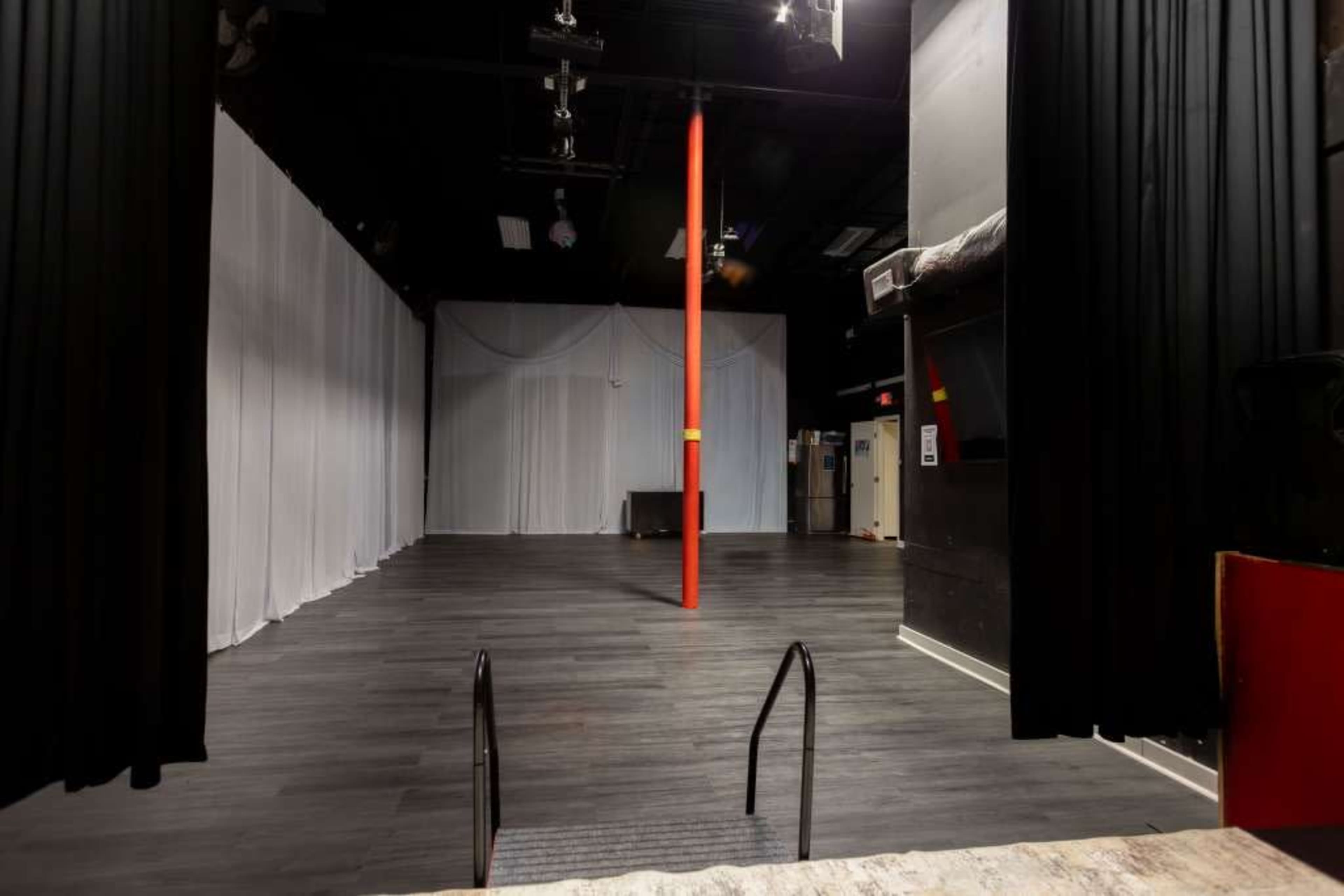 The image shows an empty stage area with a polished floor, tall black curtains, and a red pole in the center.
