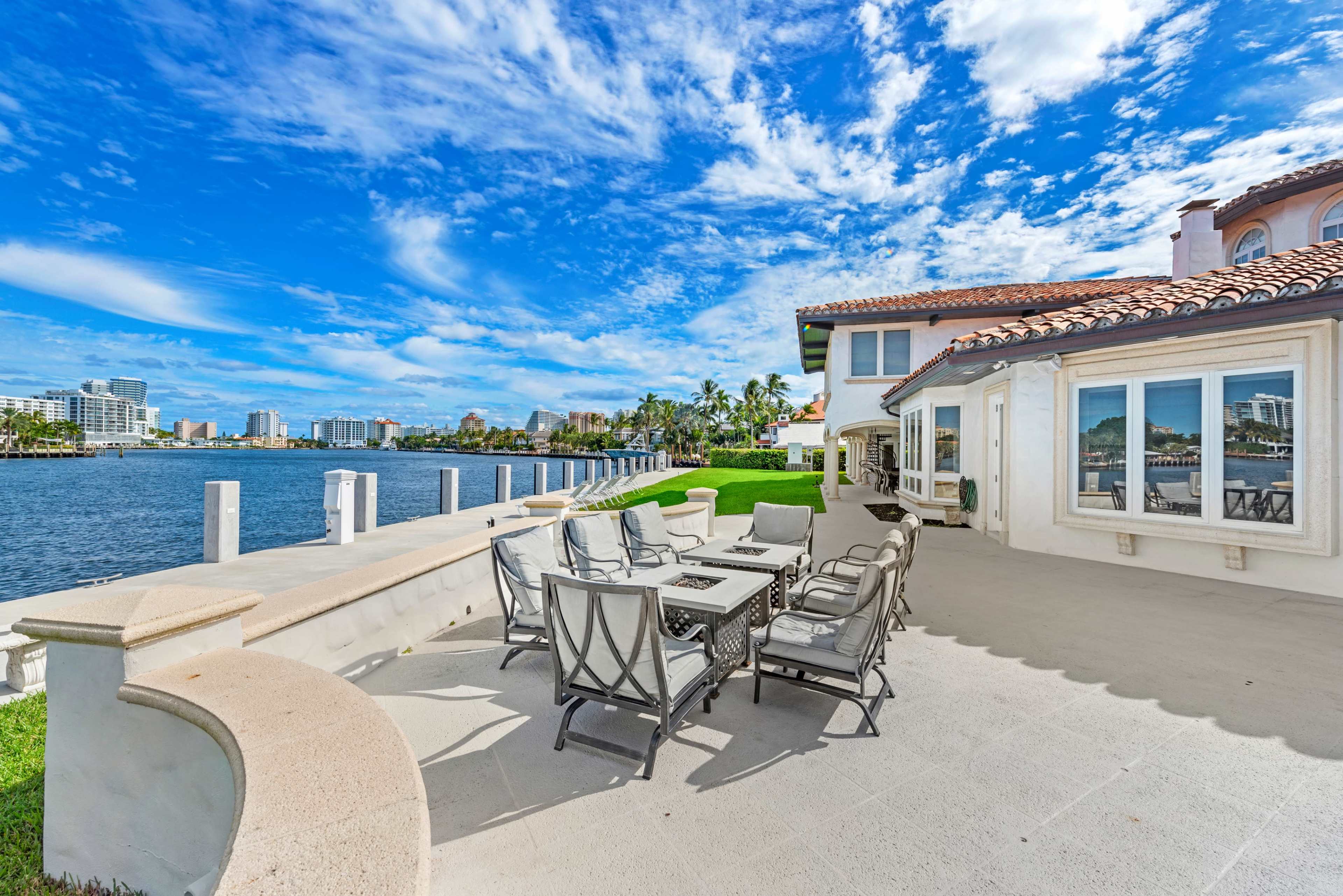 The image shows a waterfront patio with outdoor furniture overlooking a canal and city skyline.